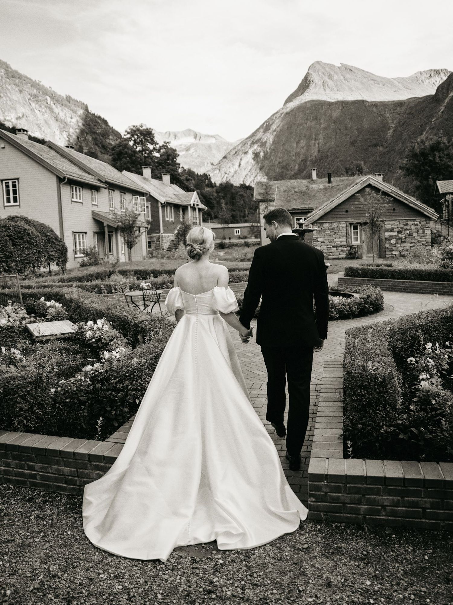 Black and white photo of a bride and groom holding hands walking through a formal garden with mountains in the background.