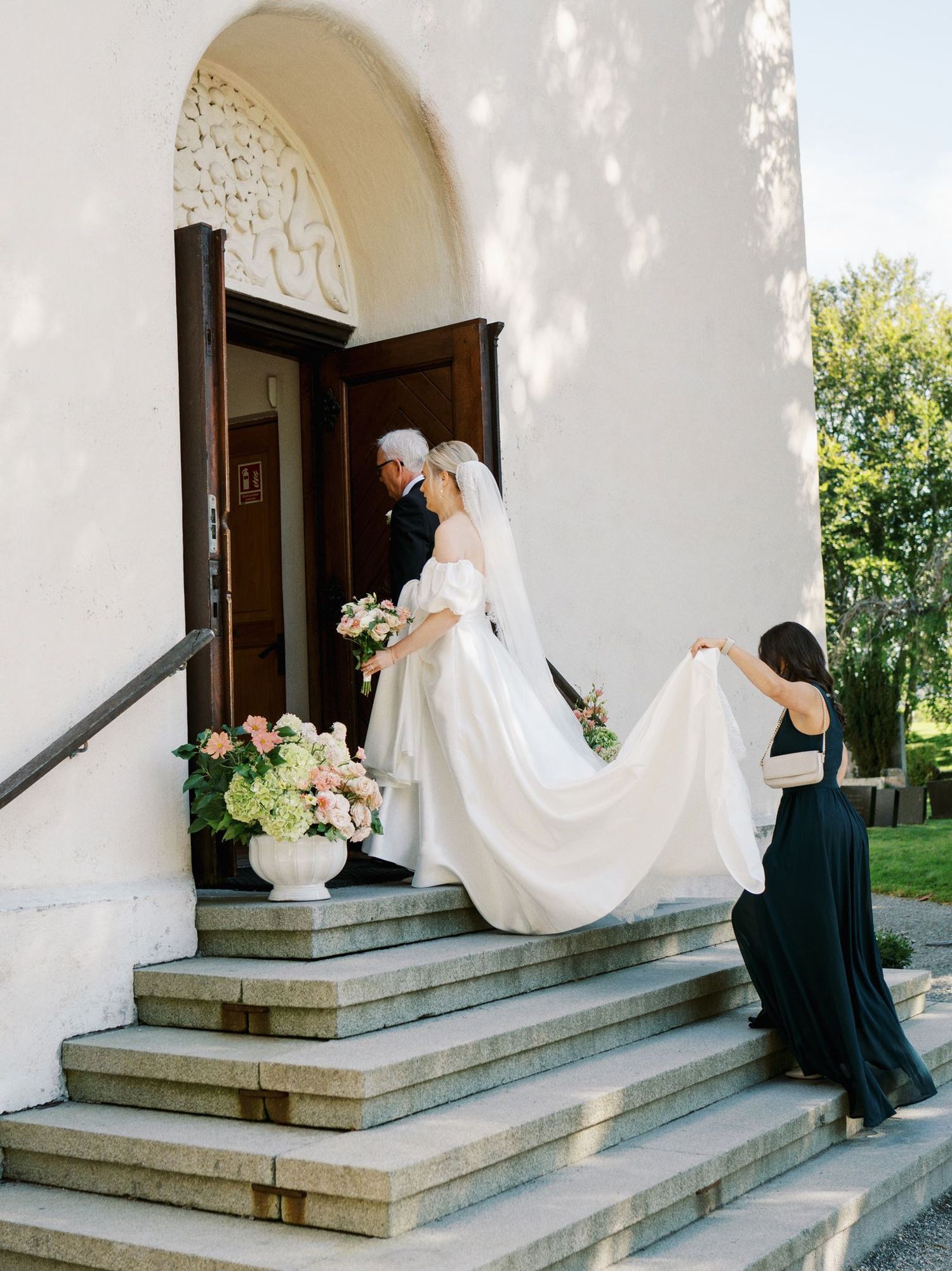 Bride ascending church steps with veil carried by bridesmaid in dark green dress, accompanied by older gentleman.
