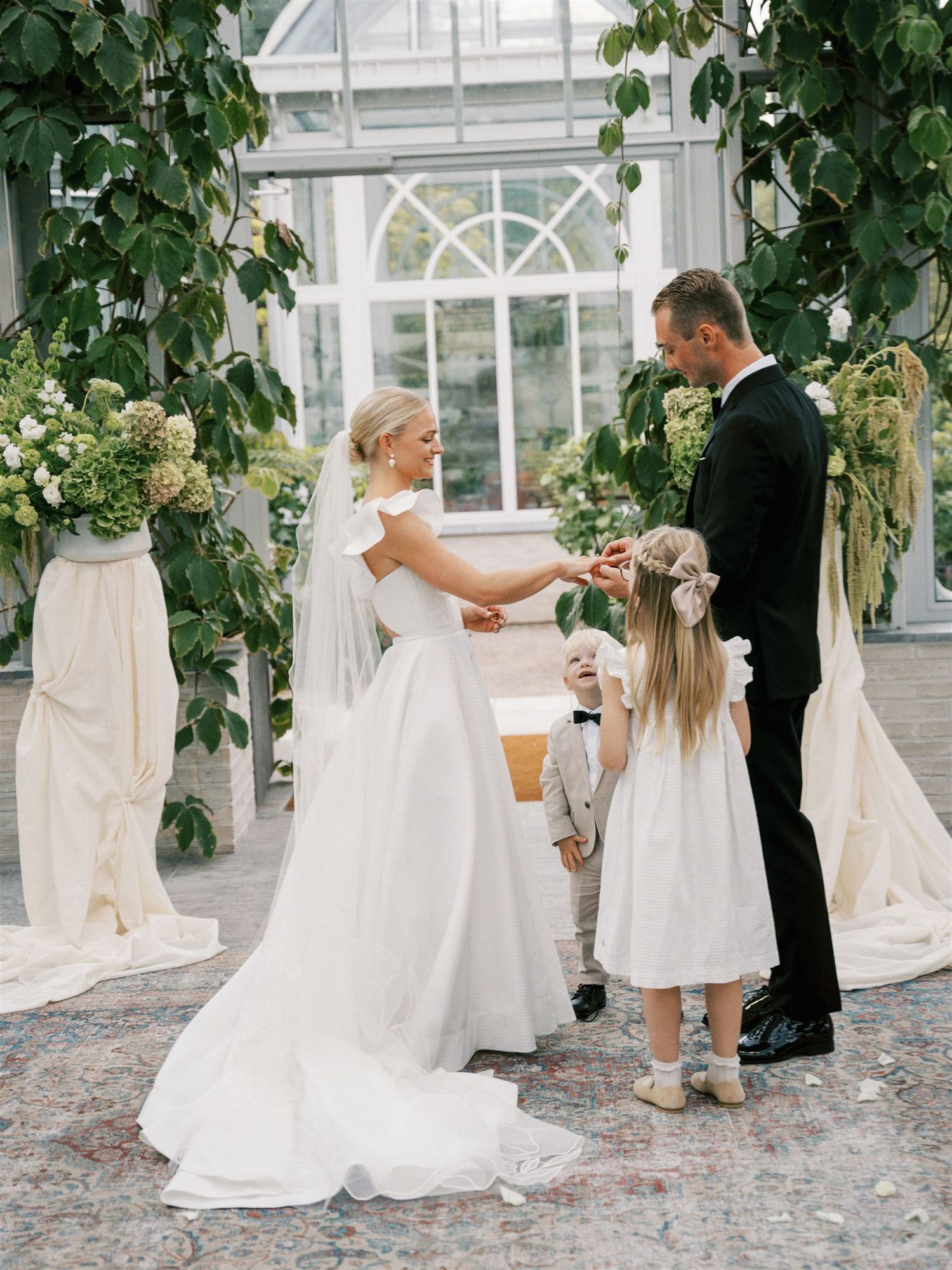 Bride and groom exchange rings at greenhouse ceremony with flower girl and lush green arch decor.