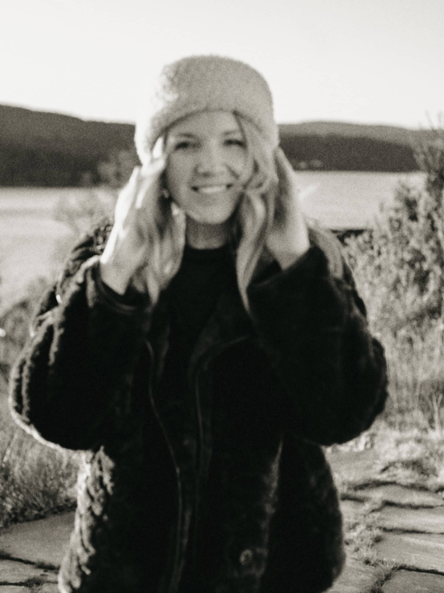 Black and white portrait of smiling blonde woman wearing knit hat outdoors near a scenic lake.