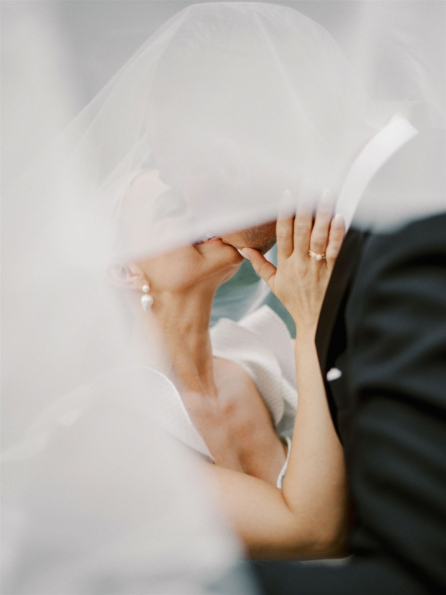 Romantic bride and groom kissing under flowing white veil in soft ethereal wedding portrait.