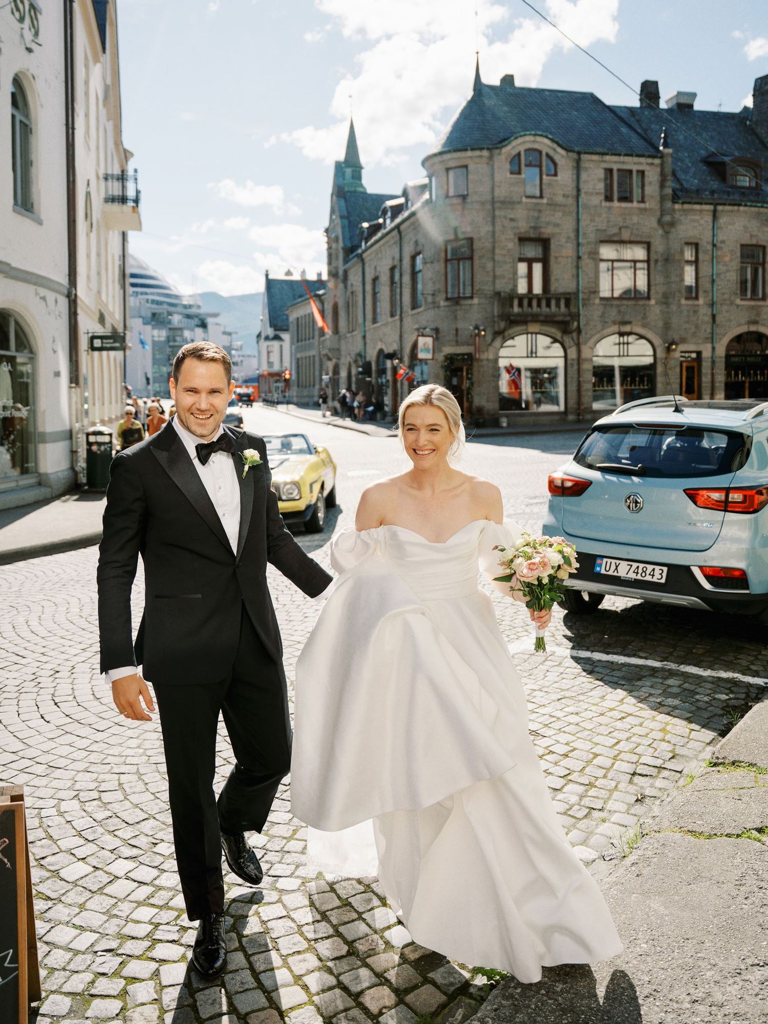 A bride and groom walk hand-in-hand on a cobblestone street in a European town, she in a white gown, he in a black tuxedo.