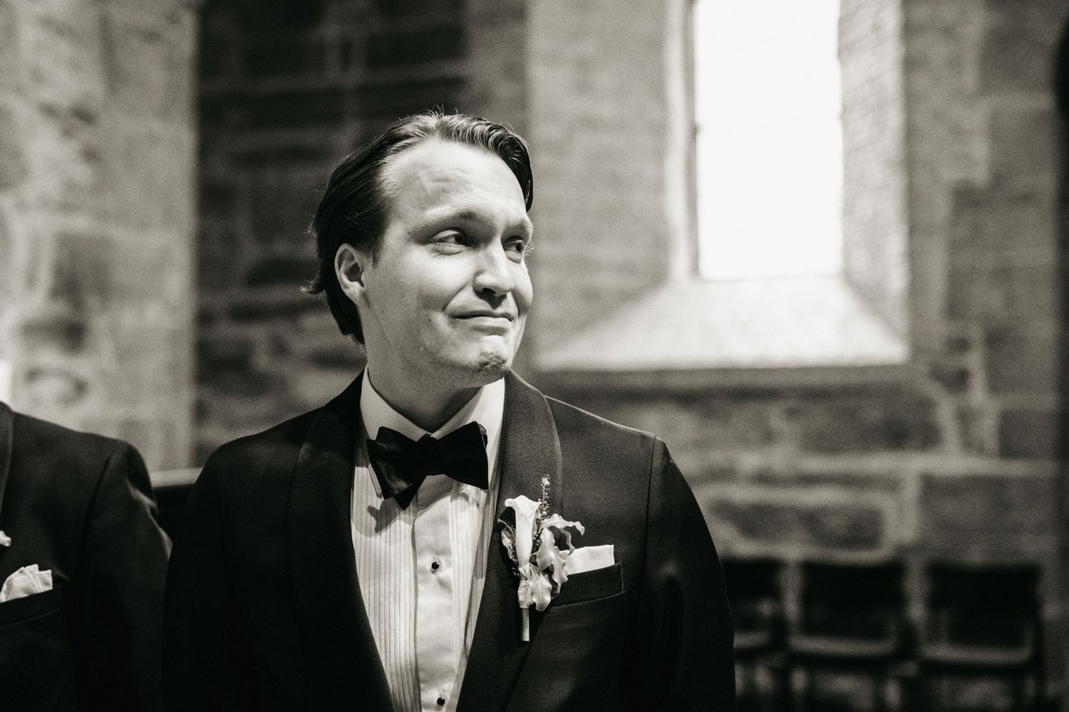 Groom in black tuxedo and bow tie smiling emotionally during wedding ceremony in stone church.