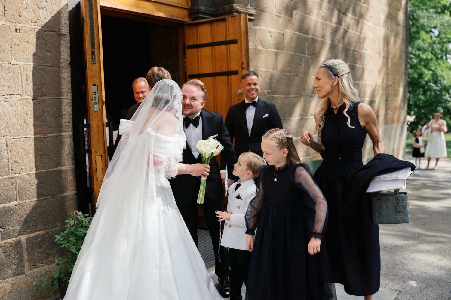 Bride in white gown and veil with groom in tuxedo greeting guests outside a stone church wedding venue.