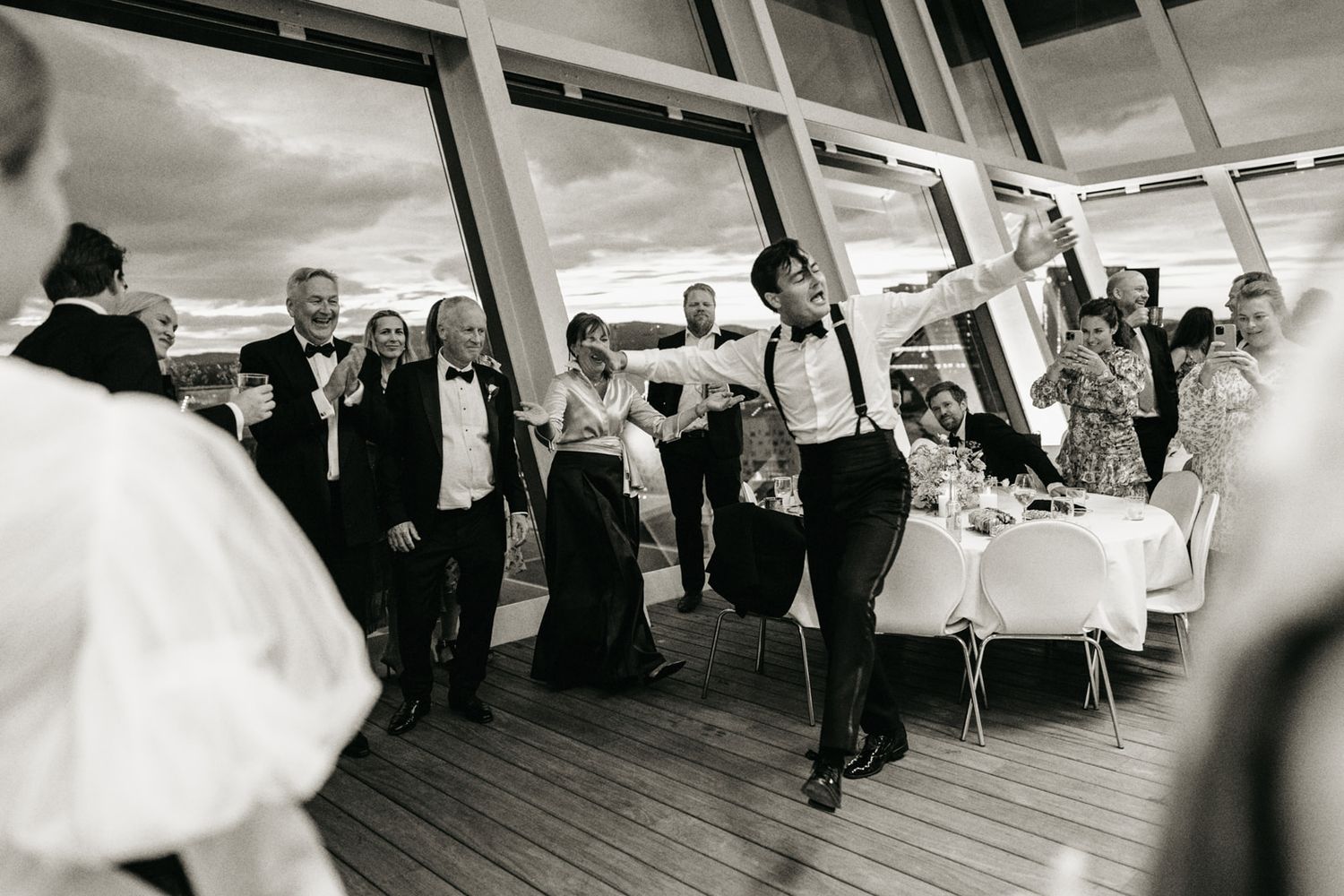 Black and white wedding reception photo with guests dancing and celebrating in an elegant venue with large windows.