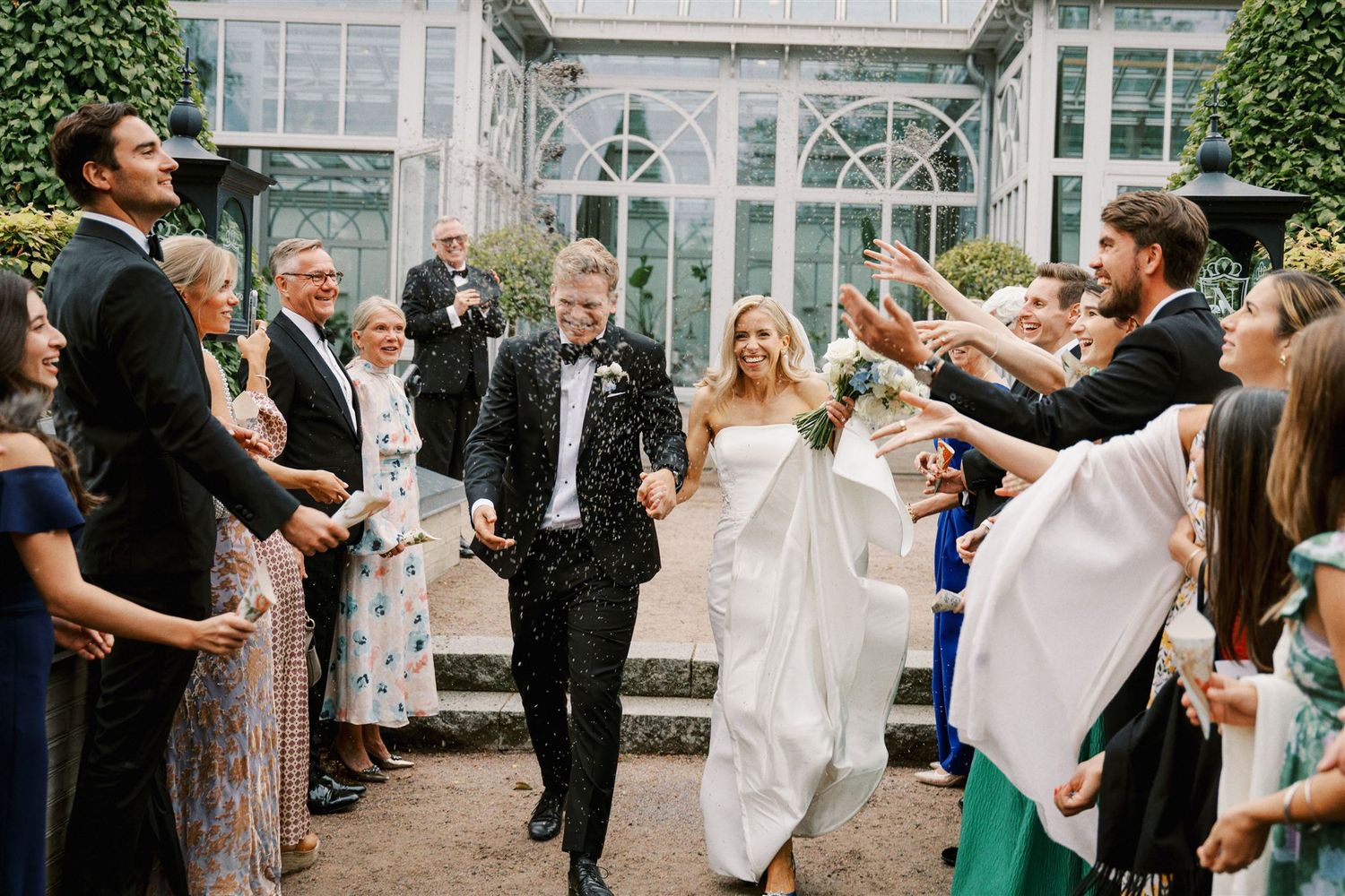Bride and groom exit wedding ceremony through confetti shower with smiling guests lining the path outside a greenhouse venue.