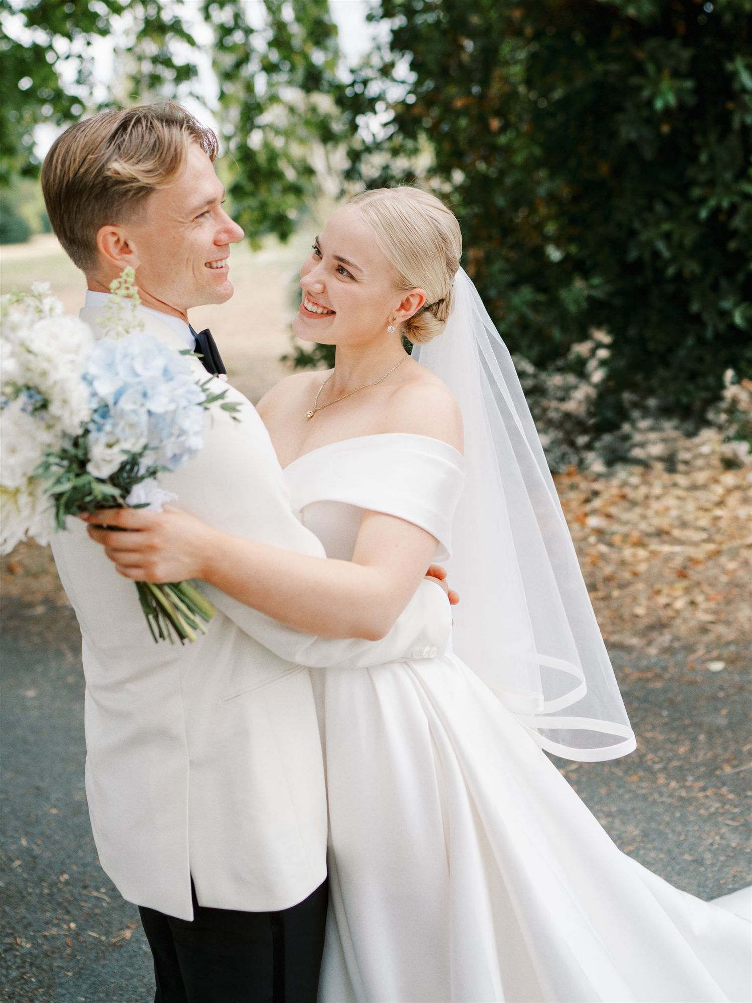 Smiling bride and groom embracing outdoors, bride in off-shoulder gown holding blue and white bouquet.