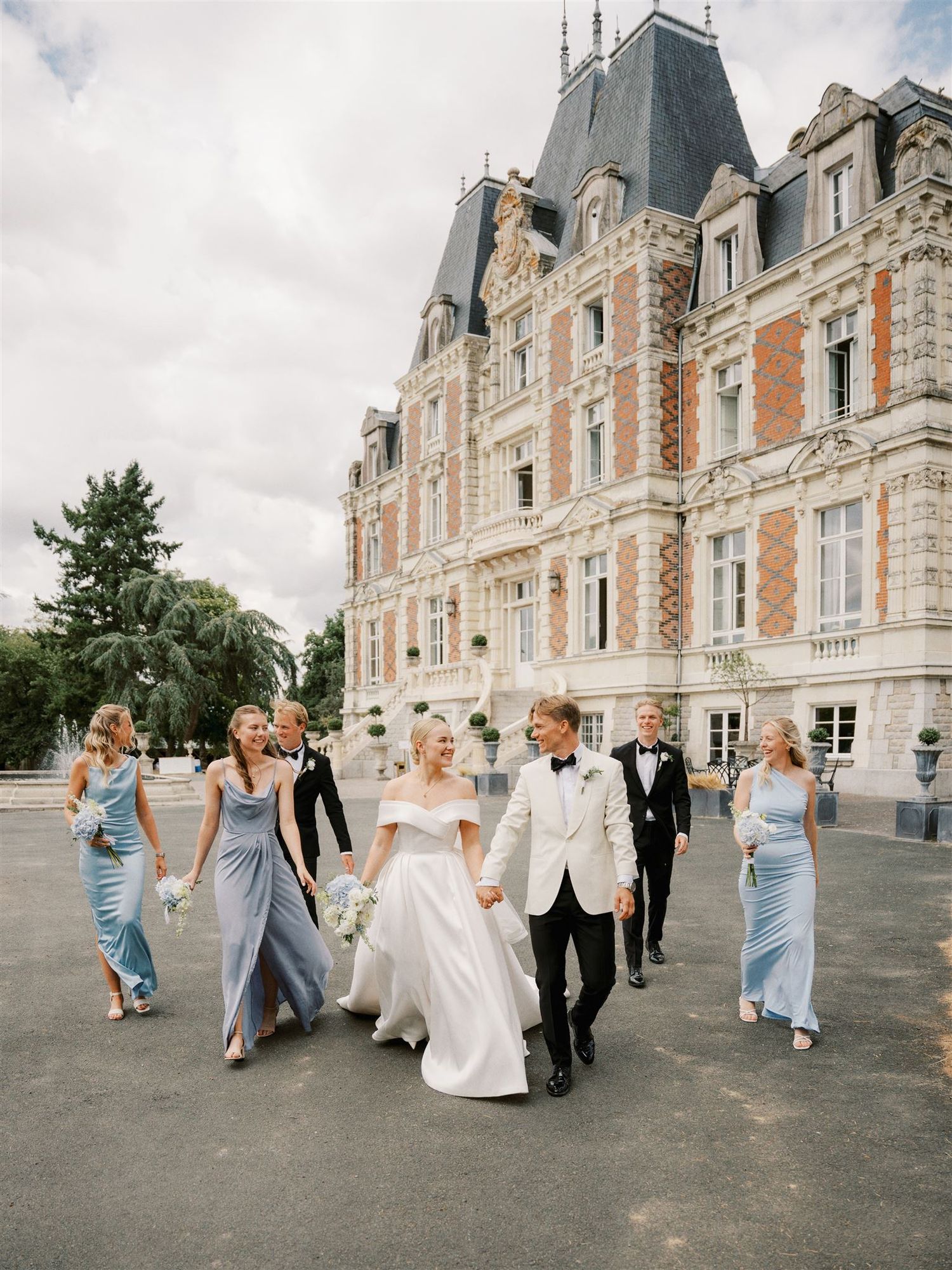 Wedding party in blue bridesmaid gowns and white tuxedo walks in front of a grand French chateau.