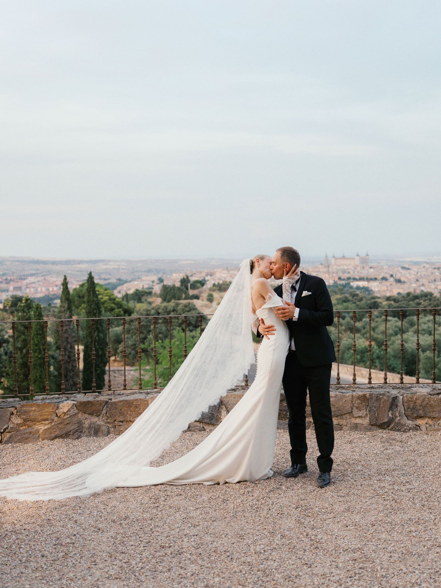 Bride and groom kissing at scenic overlook, bride in fitted gown with cathedral veil, groom in black tuxedo.