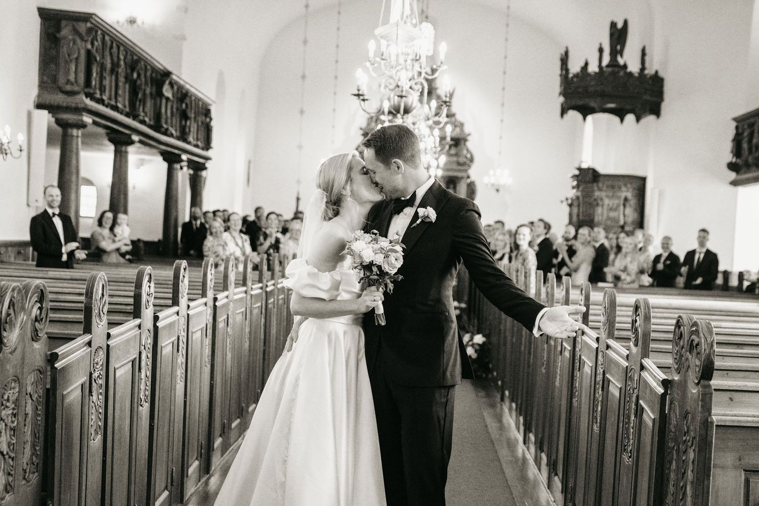 Black and white photo of a bride and groom sharing a kiss in a church aisle after their wedding ceremony.