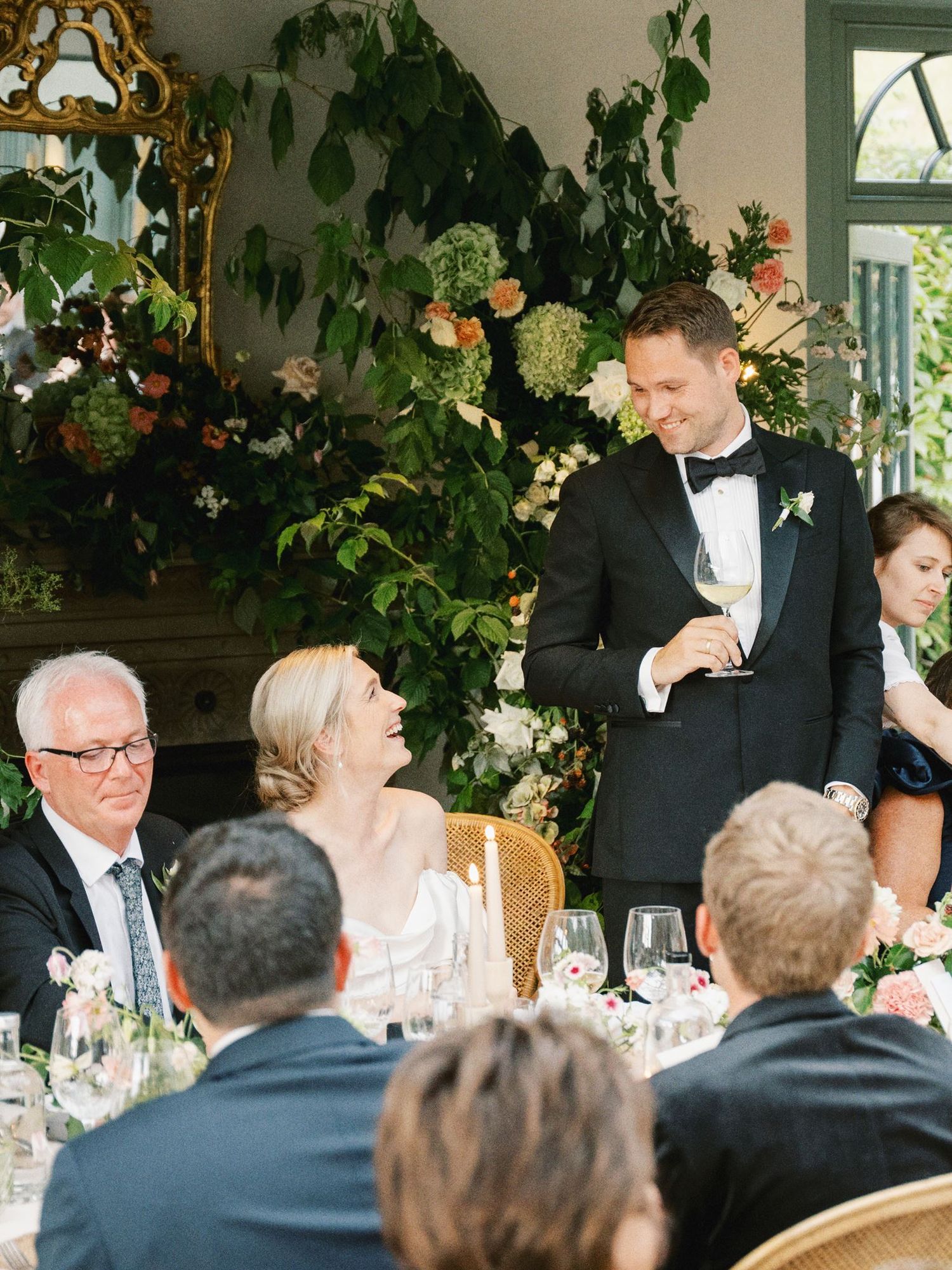 Groom in black tuxedo giving a wedding toast while guests smile and applaud at an elegant floral reception.