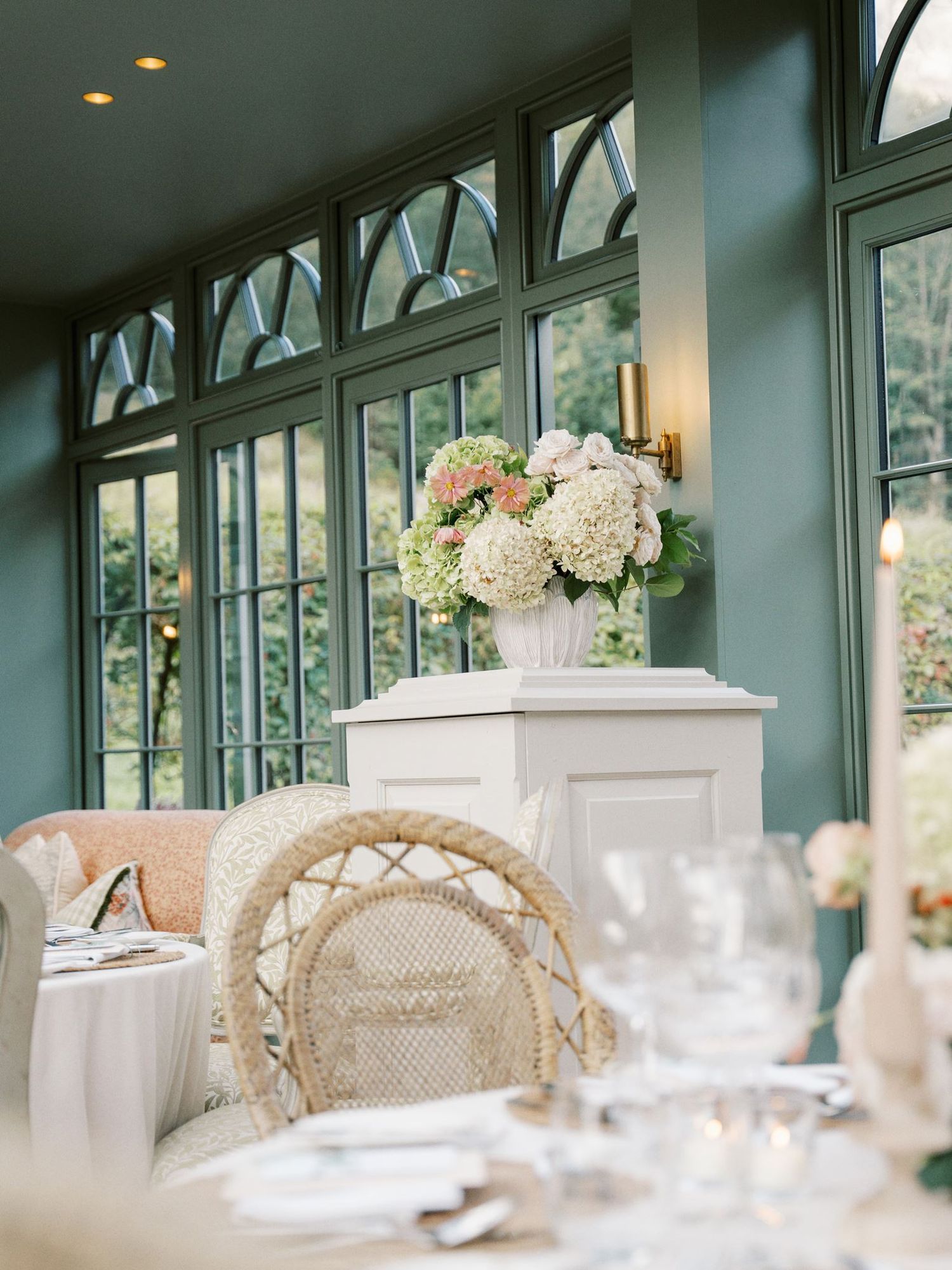 Elegant wedding reception in a green conservatory with wicker chairs, white hydrangea floral arrangement, and candlelit dining tables.