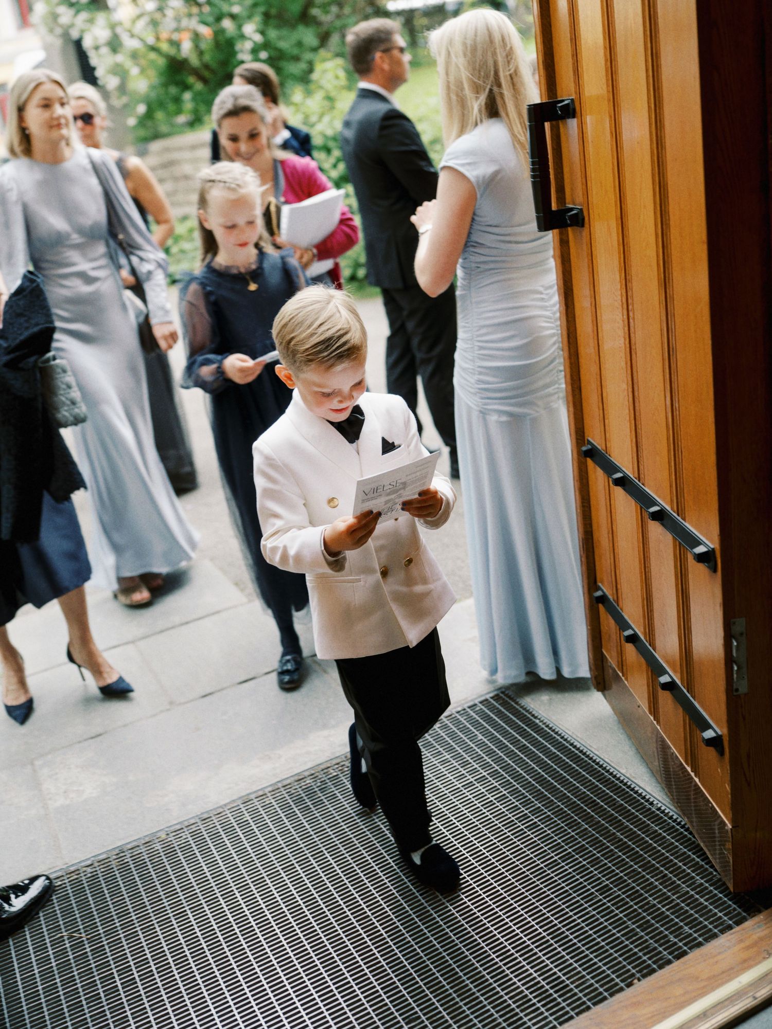 Young ring bearer in white tuxedo jacket reads wedding program at church entrance with guests in background.