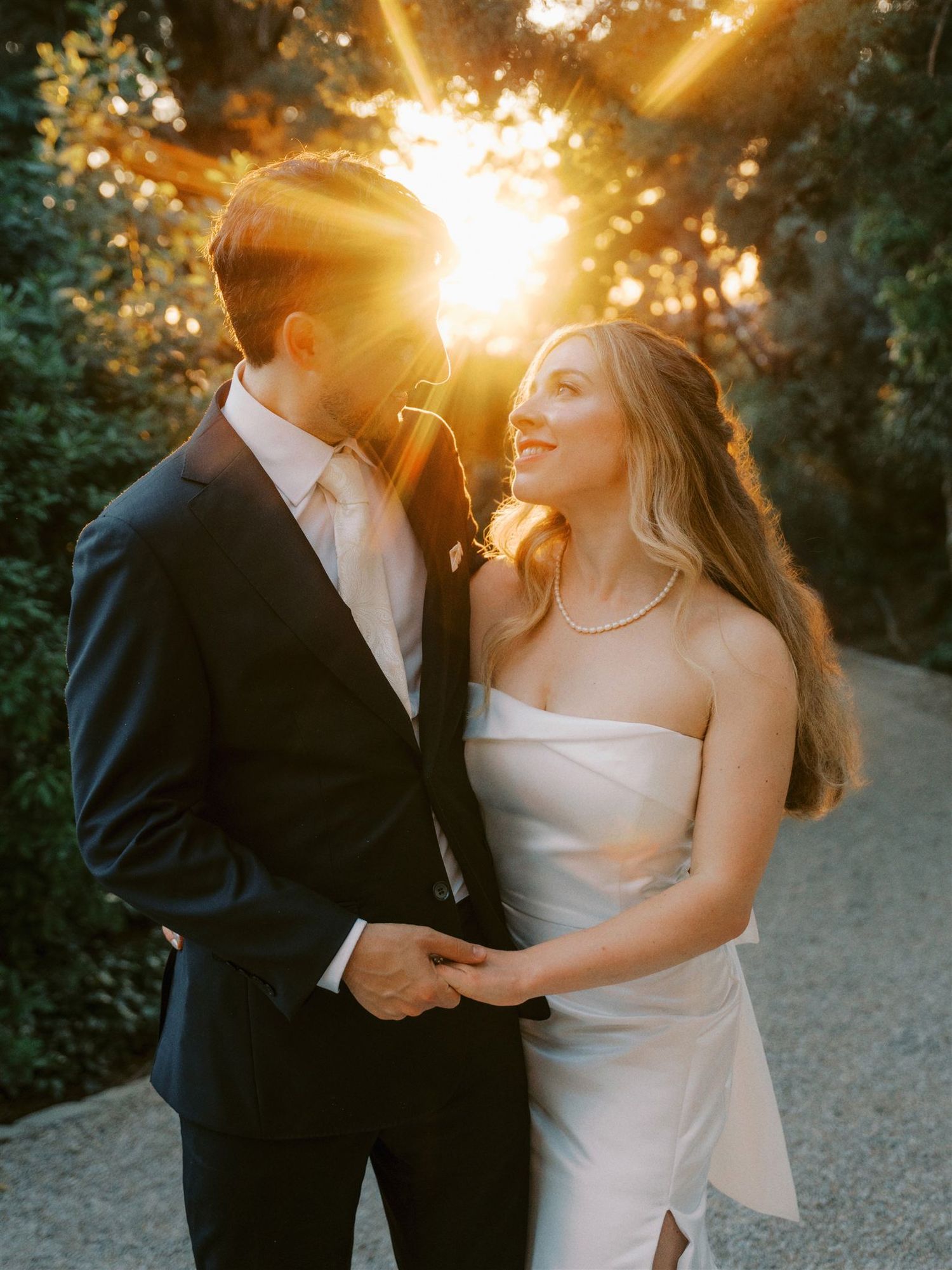Bride and groom sharing a romantic moment bathed in golden sunburst light among lush green trees.