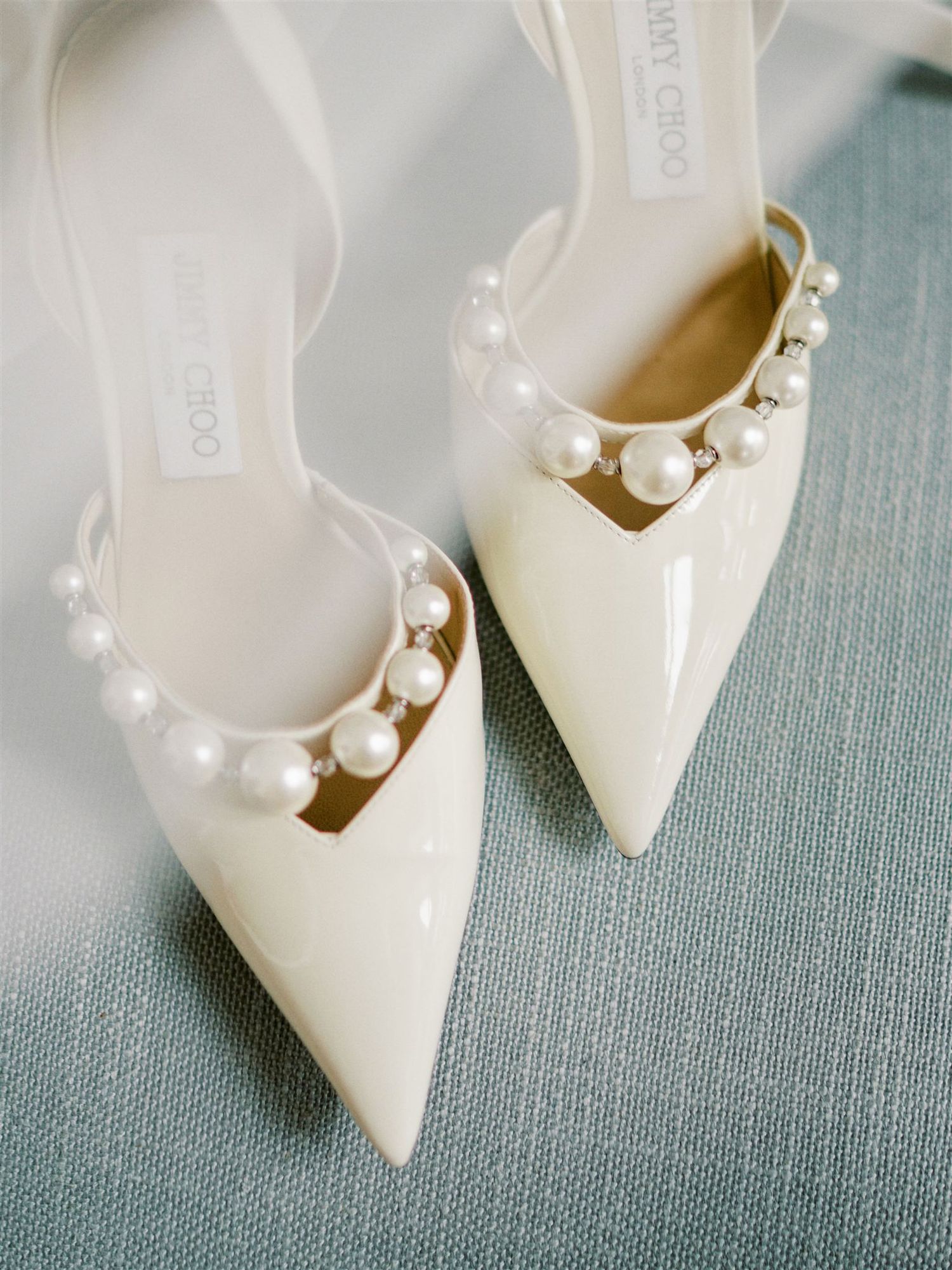 Jimmy Choo ivory patent leather pointed toe heels adorned with pearl embellishments on blue fabric.