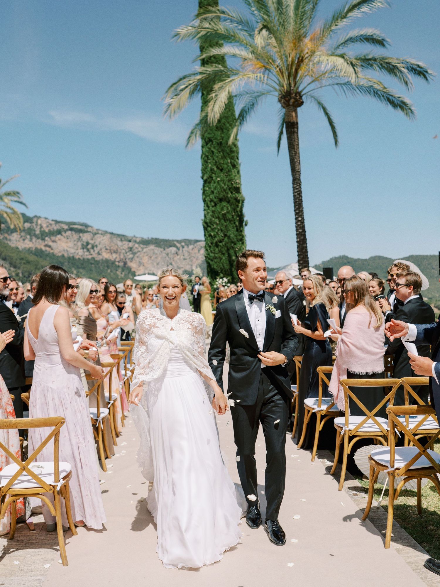 Bride and groom walk down outdoor wedding aisle lined with guests, palm trees, and mountains in background.