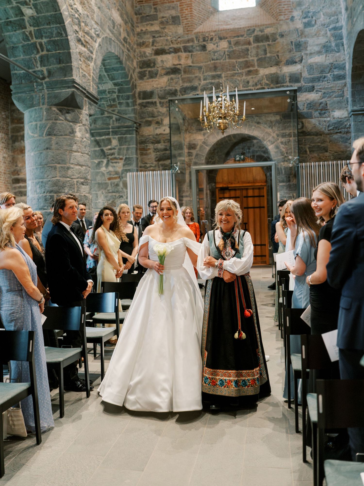 Bride in white gown walks down stone church aisle with woman in traditional Norwegian bunad dress during wedding ceremony.