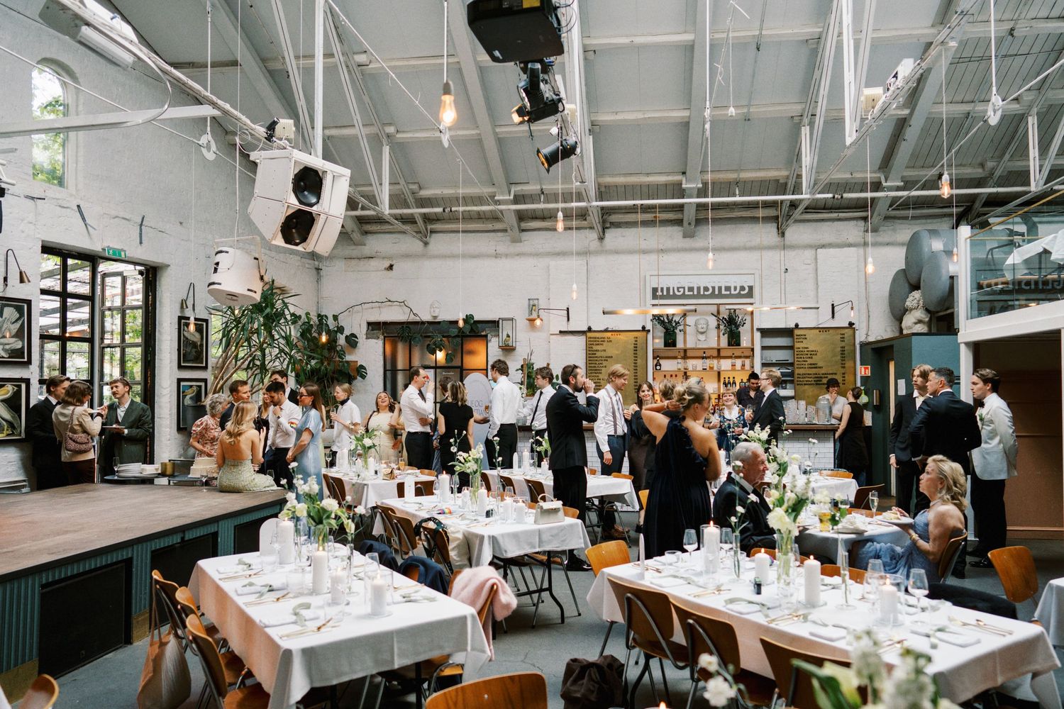 Guests mingling at a modern industrial wedding reception venue with white tablecloths and green floral decor.