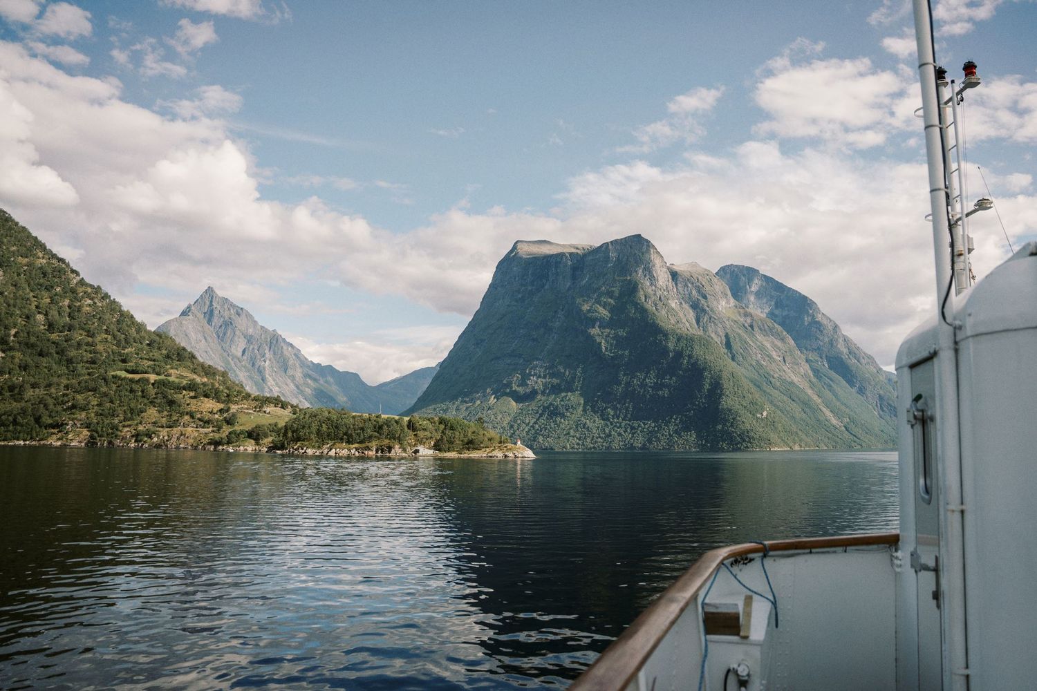 Scenic fjord view from a boat deck with calm waters reflecting dramatic Norwegian mountains under a partly cloudy sky.