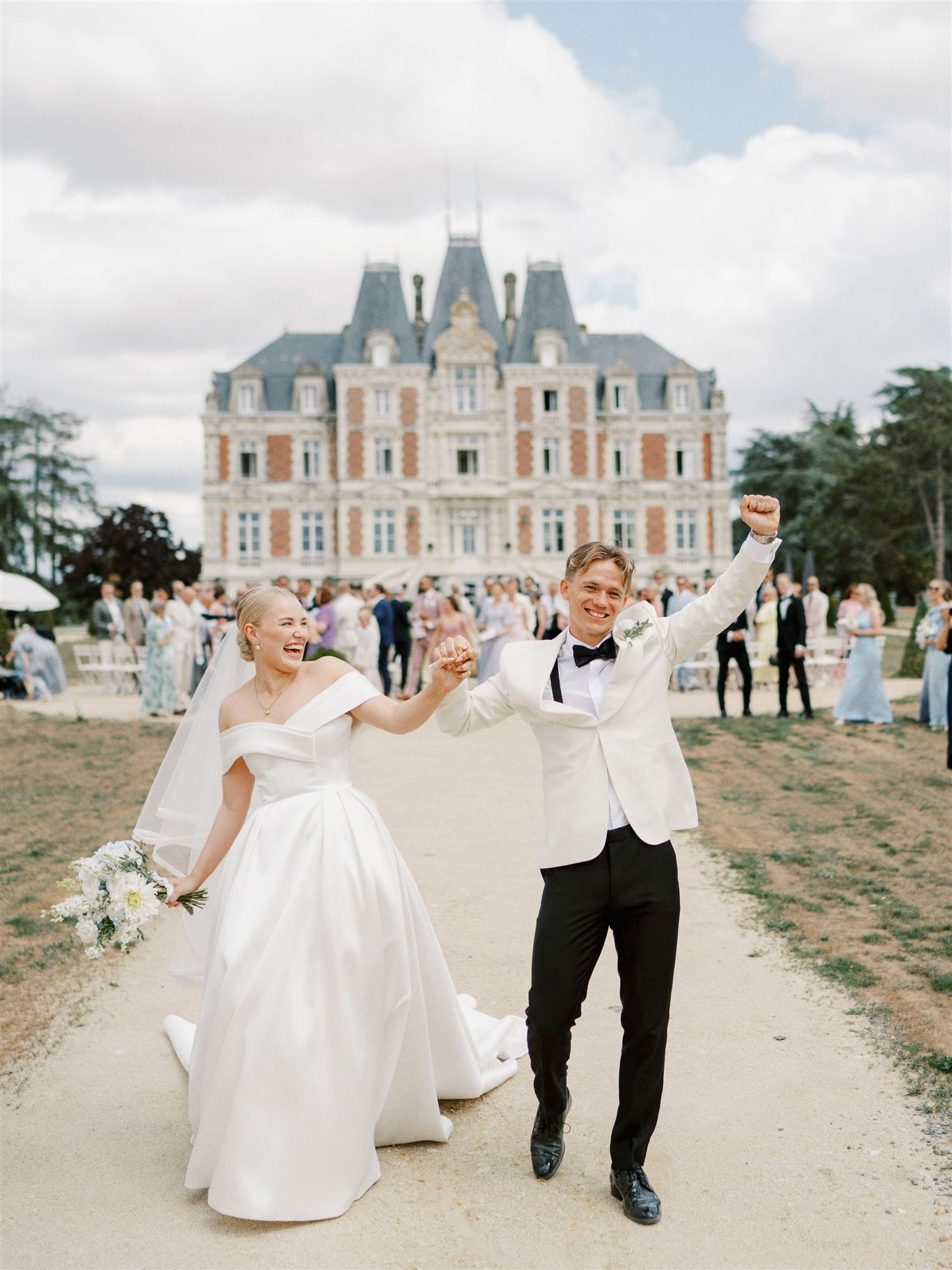 Joyful bride and groom celebrating outside a grand French chateau wedding venue with guests in background.
