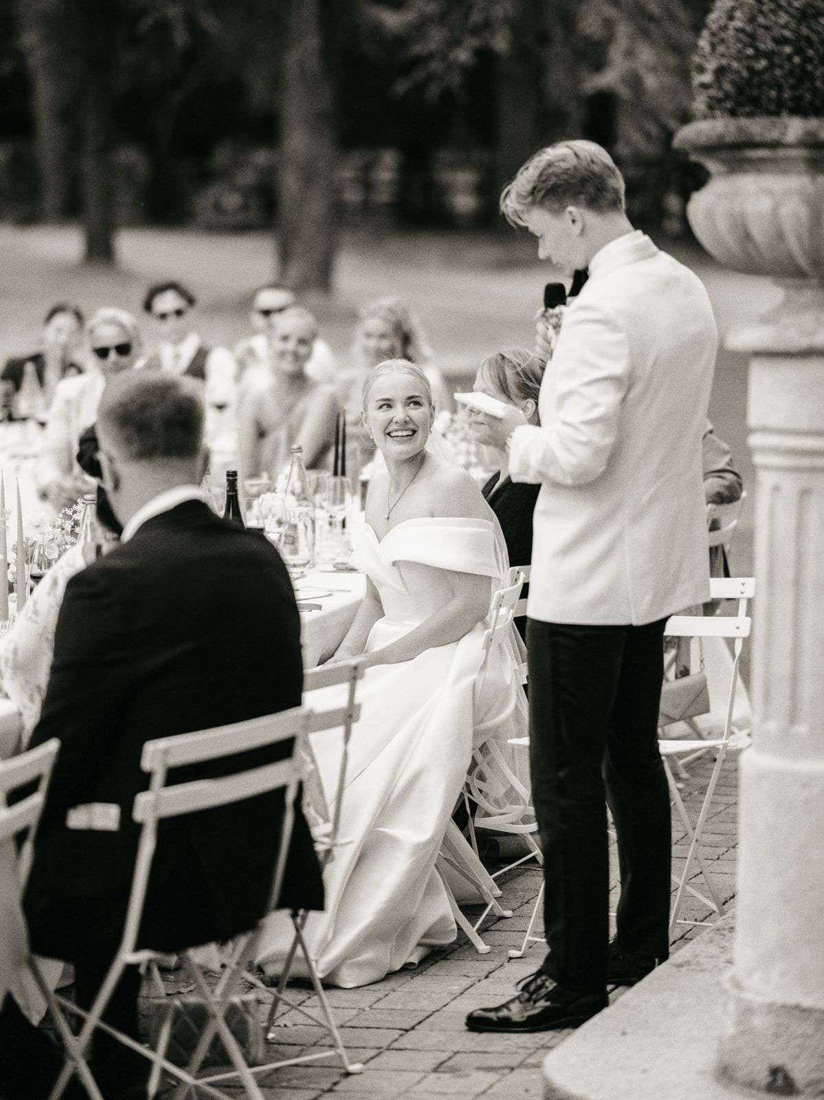 Black and white photo of groom giving a toast while bride smiles at outdoor wedding reception.