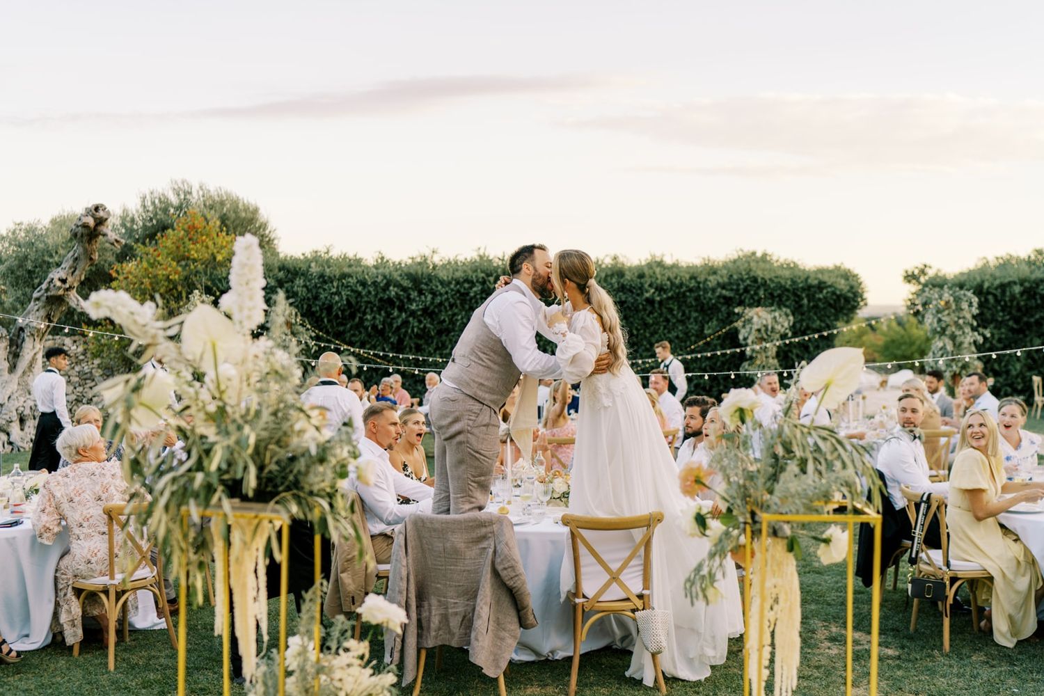Couple shares first kiss at outdoor boho reception with pampas grass, gold chairs, and string lights.