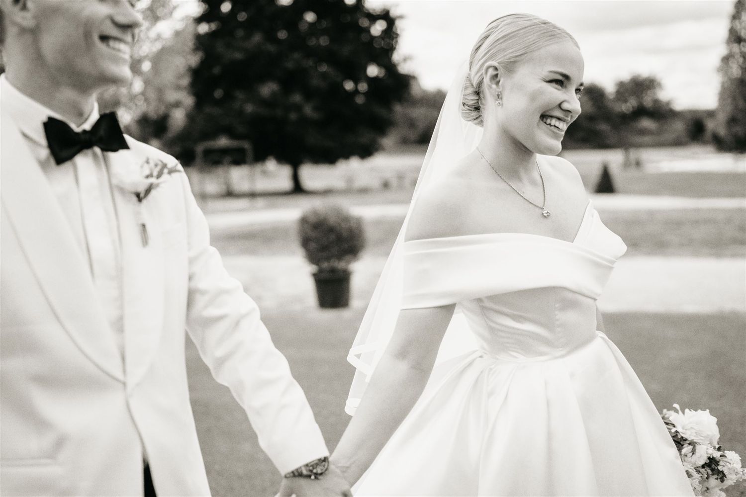 Smiling bride in off-shoulder gown and veil walking with groom in white tuxedo jacket.