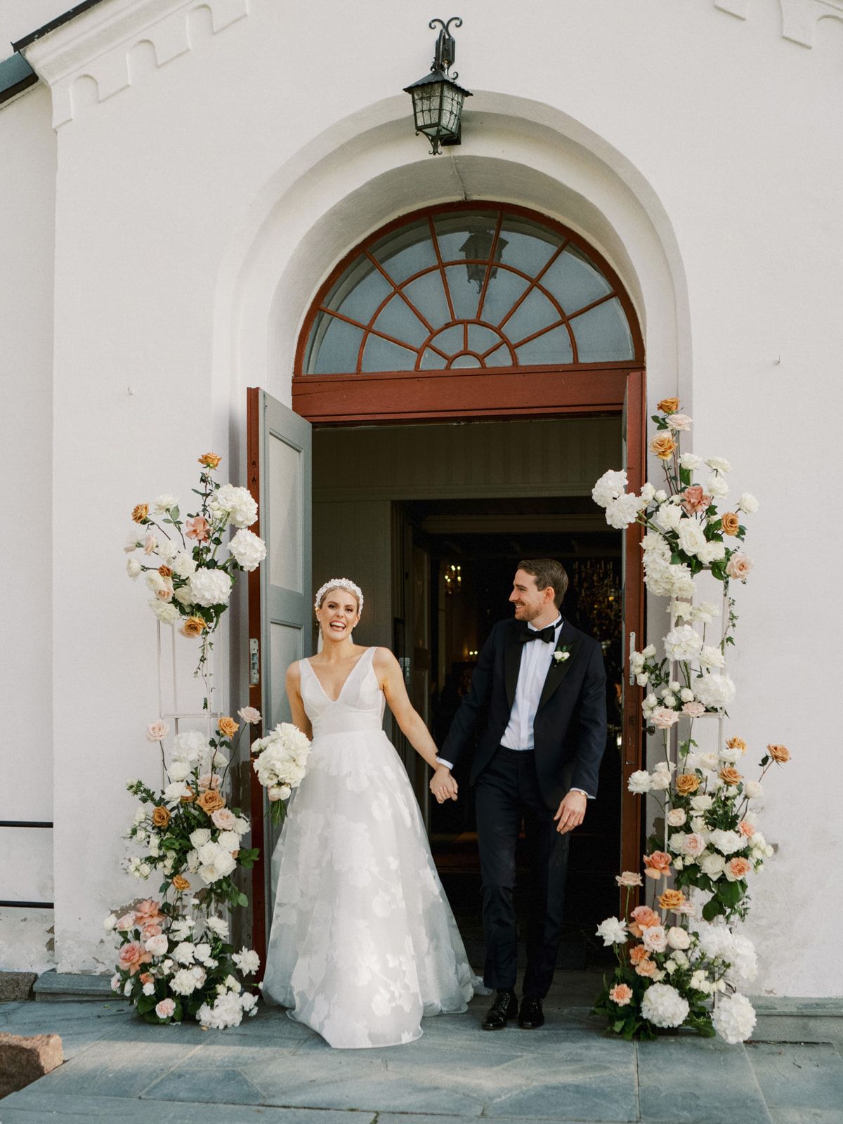 Bride and groom exit white church doorway framed by peach and white floral arrangements.