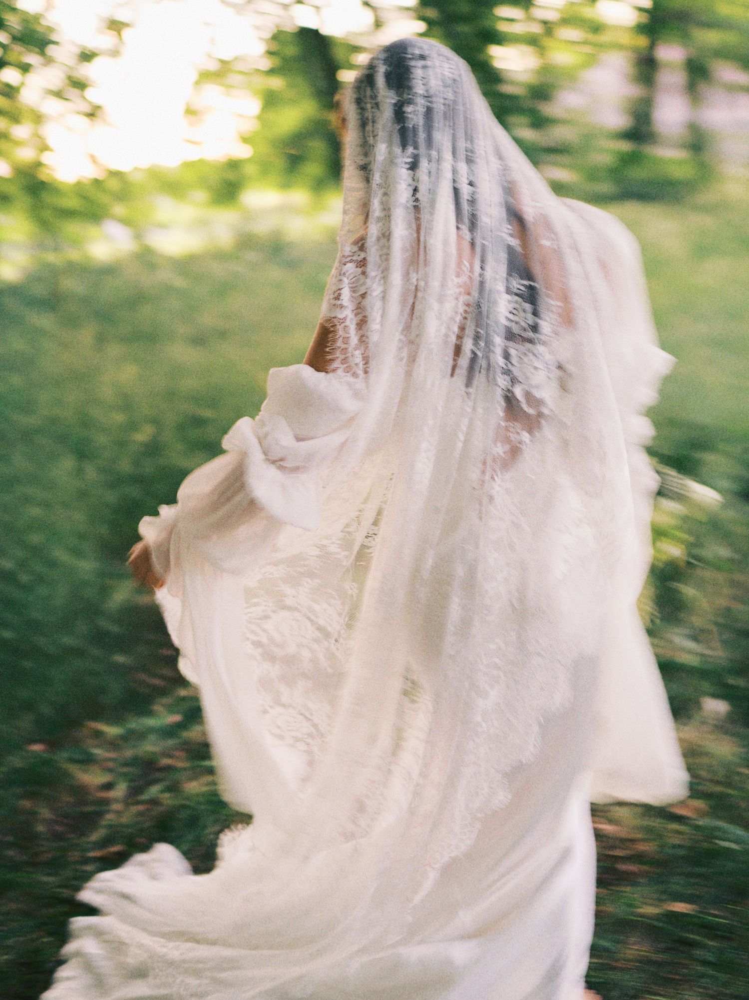 Motion blur photo of bride running through green forest in lace wedding gown and cathedral veil.