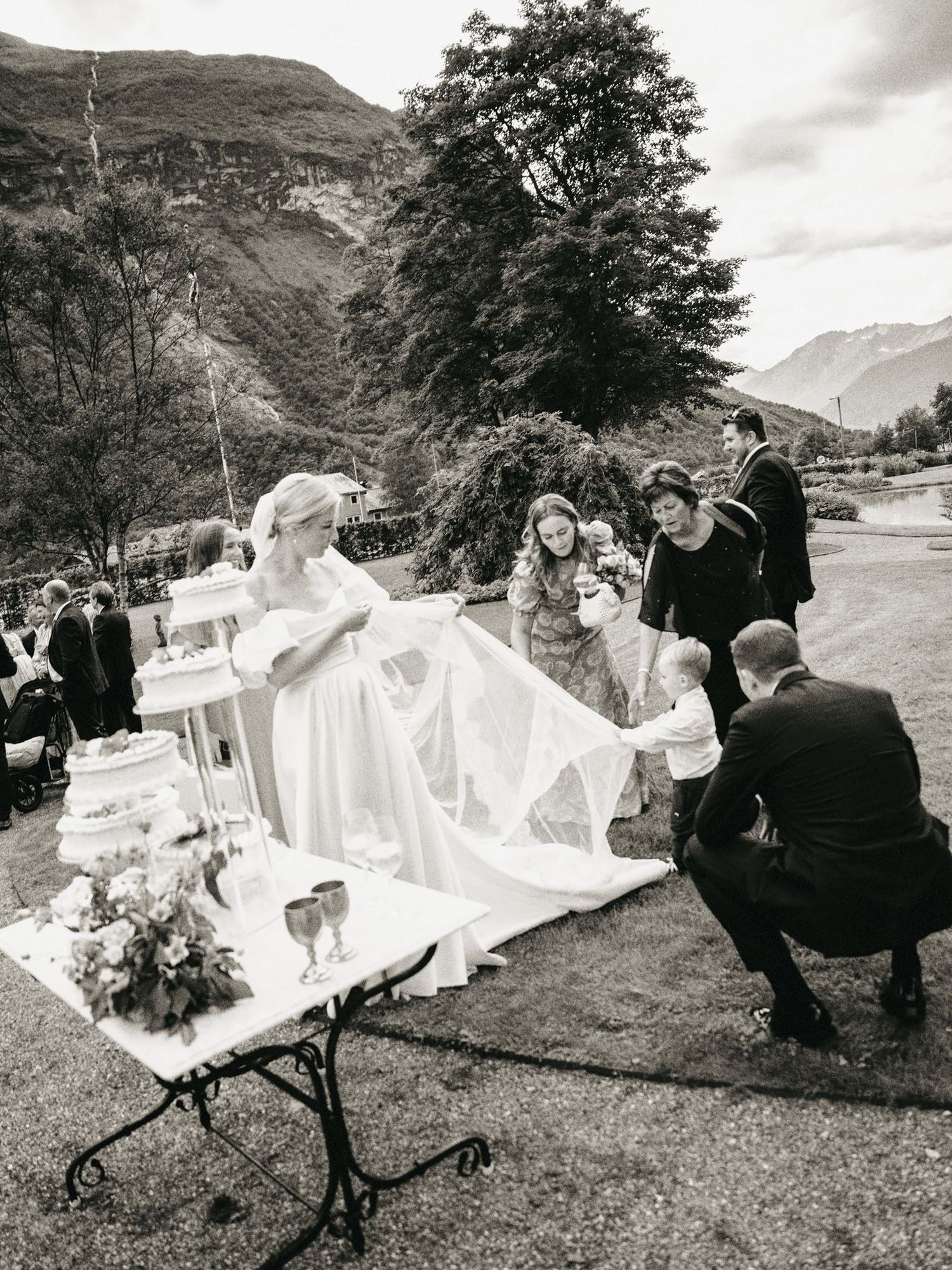 Black and white outdoor wedding ceremony with bride in gown, guests, and mountain backdrop.