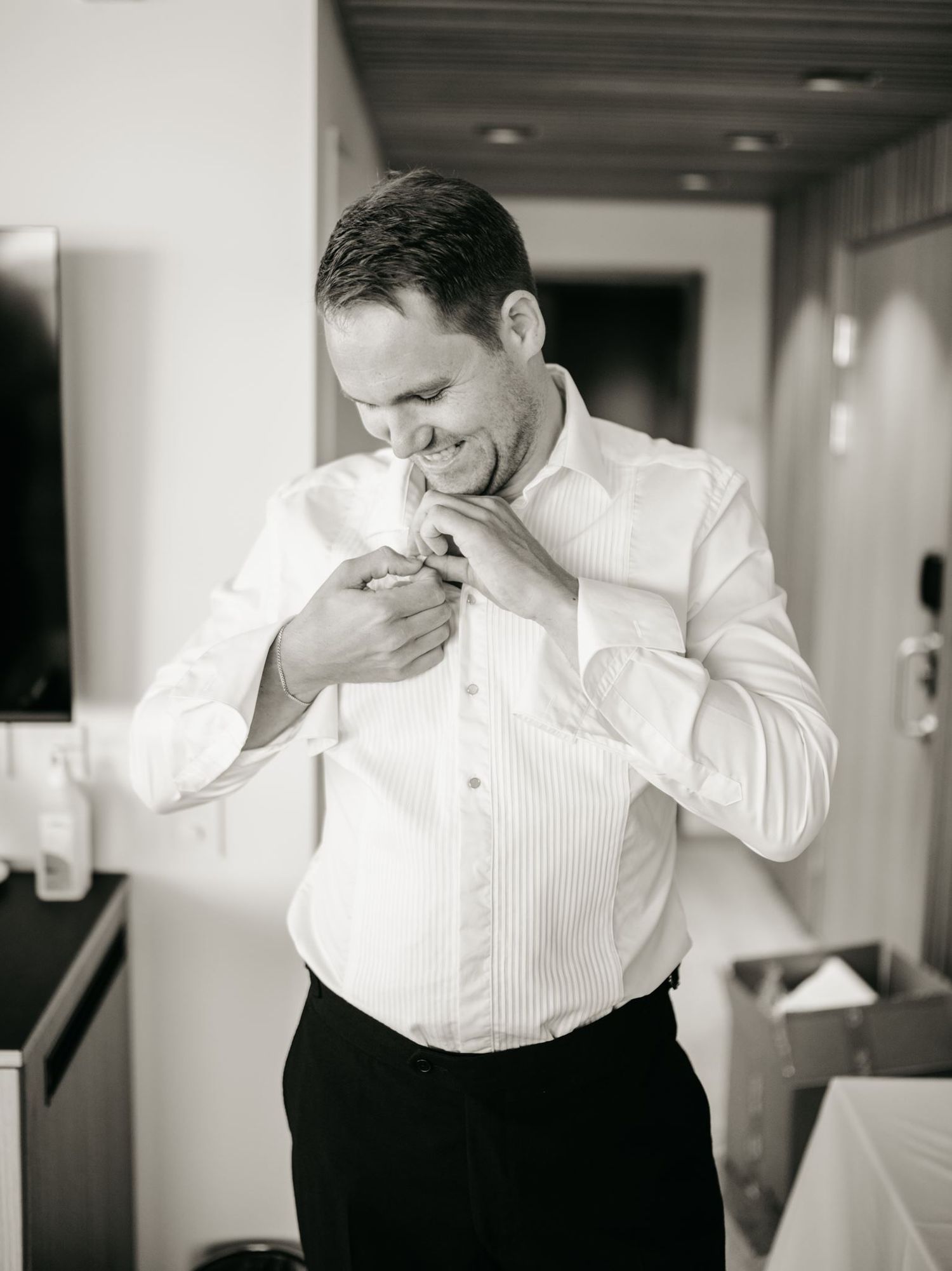Groom in white dress shirt adjusting his collar while getting ready for wedding ceremony in black and white photo.