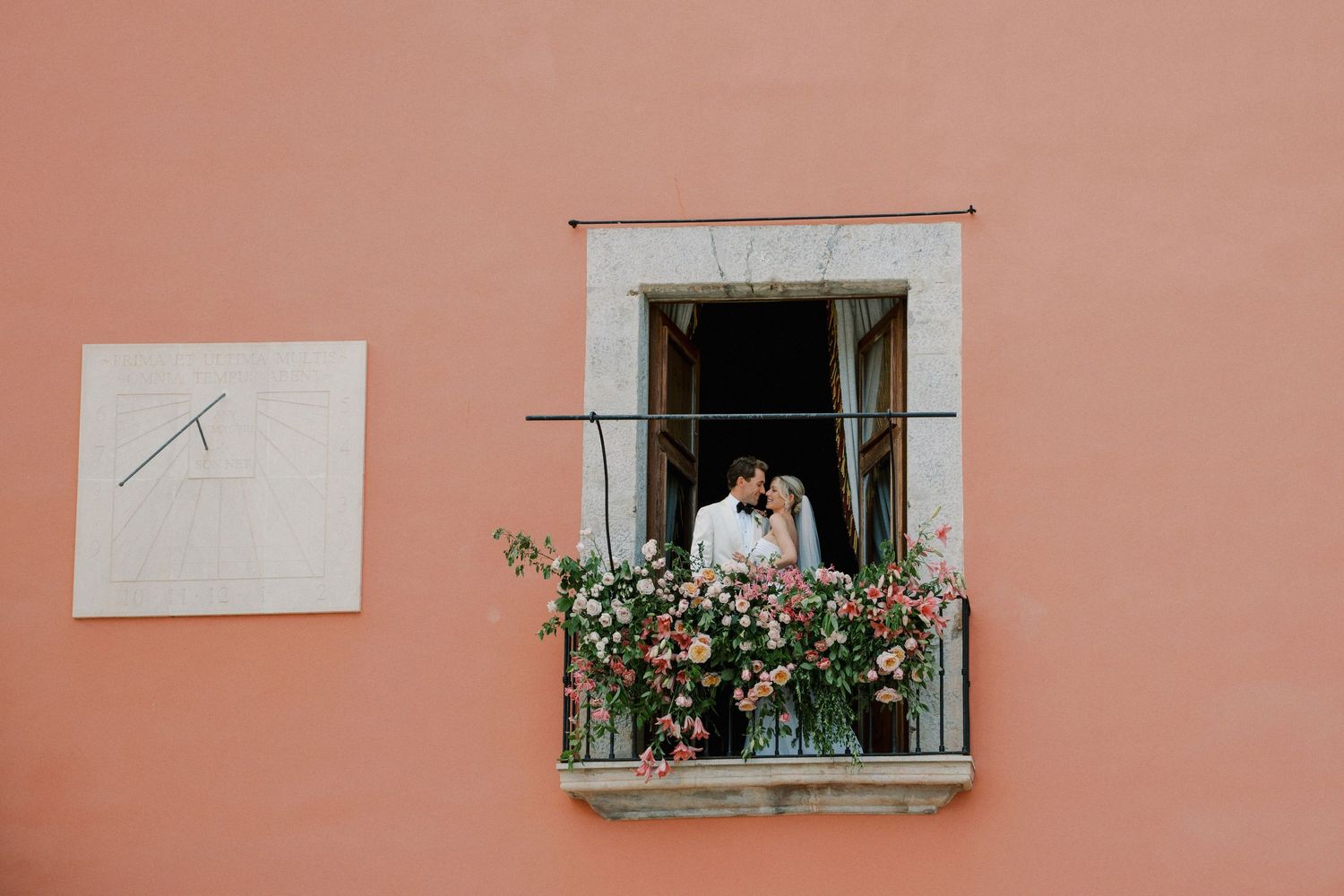 Couple kissing on a flower-adorned balcony of a pink building with a sundial nearby.