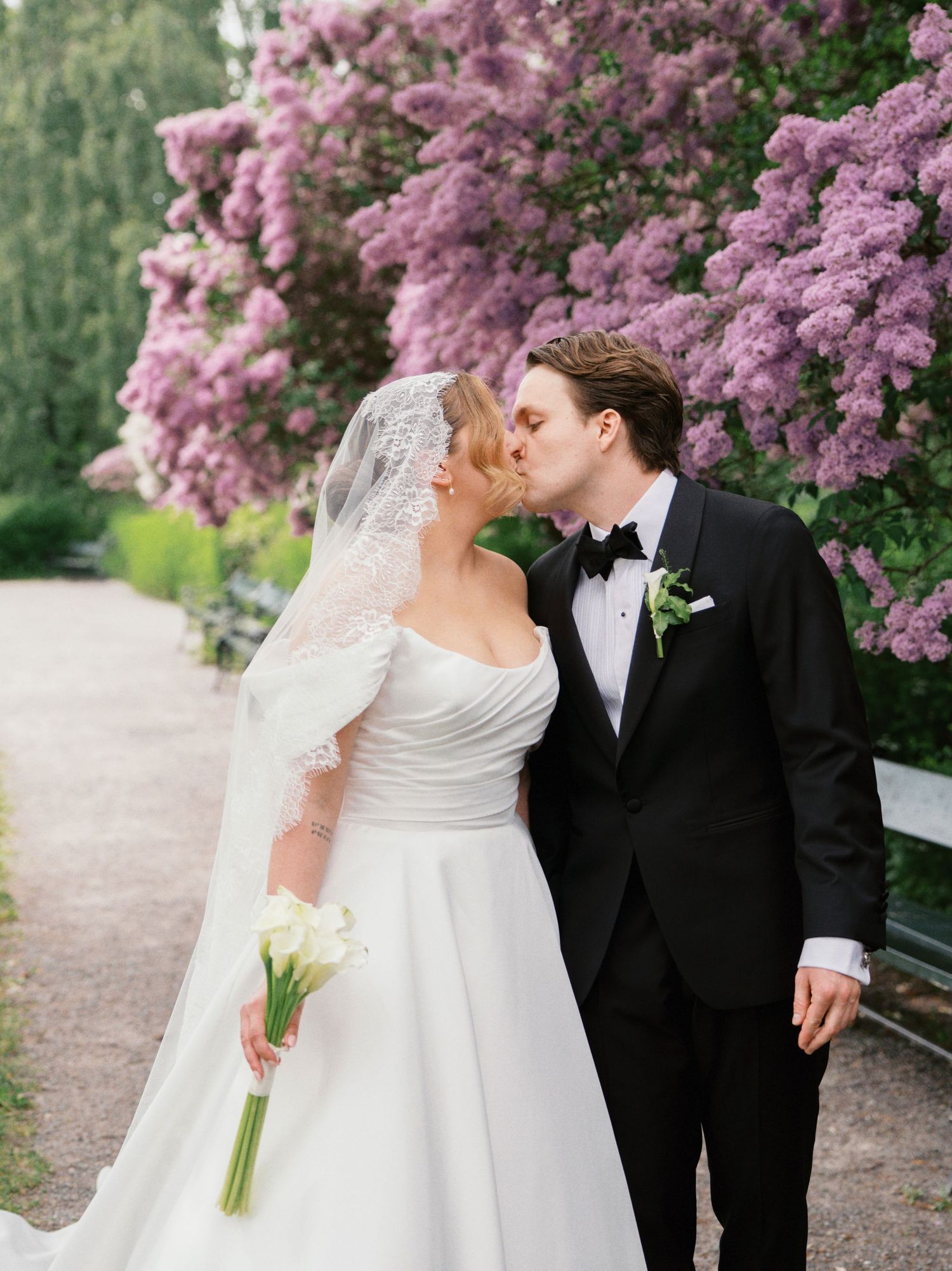 Bride and groom kissing in front of blooming purple lilac bushes, bride holding white calla lily bouquet.