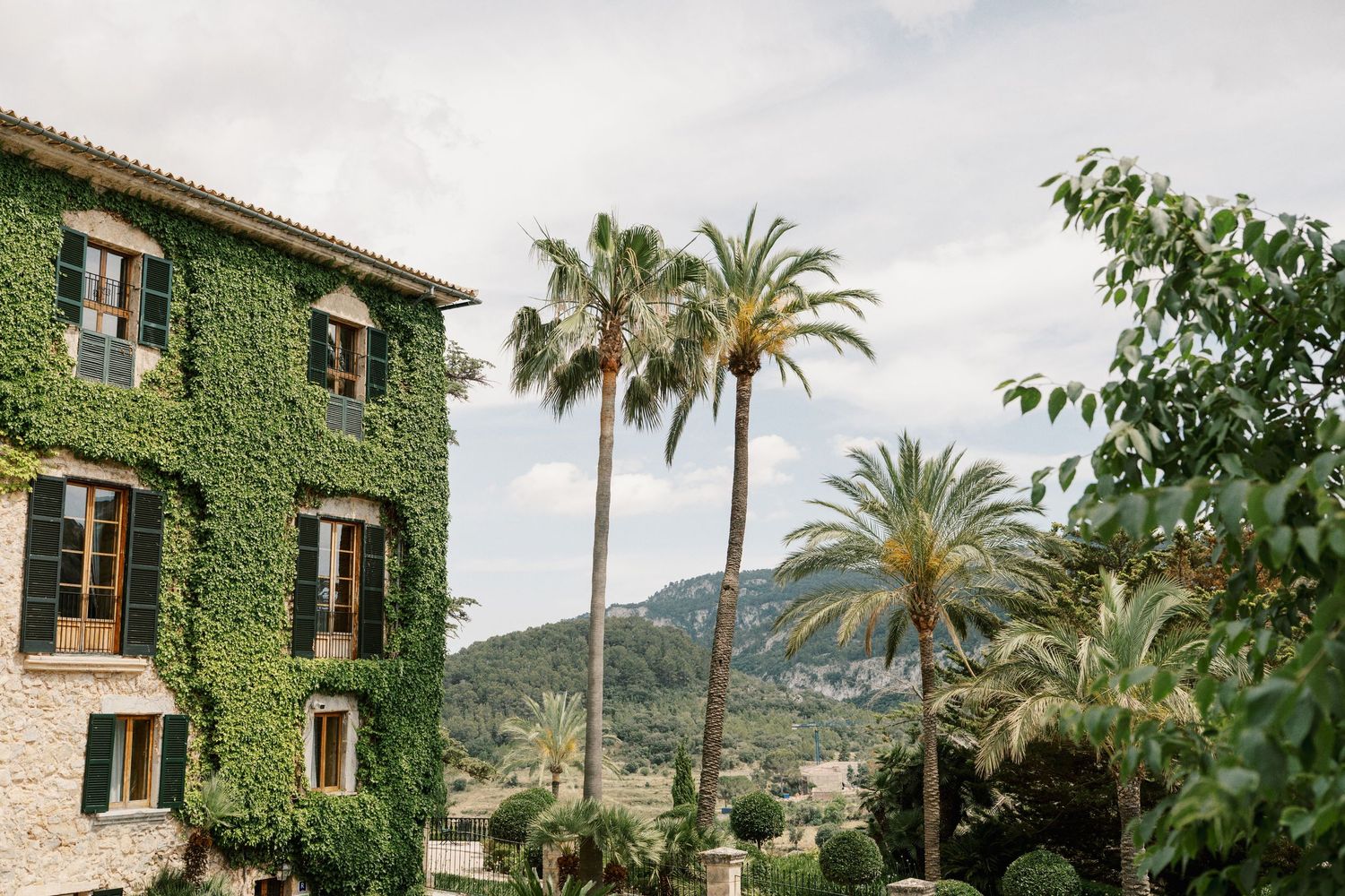 Ivy-covered stone building beside tall palm trees with mountain views under a cloudy sky in Mallorca, Spain.