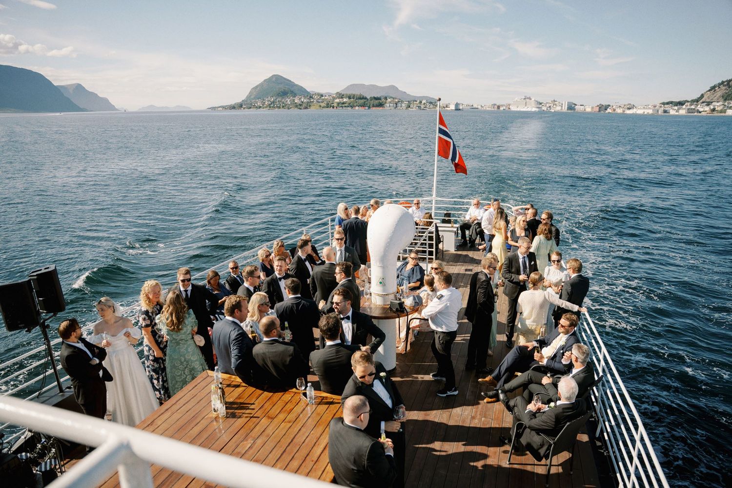 Outdoor wedding ceremony on a boat deck in Norway, guests seated around couple, Norwegian flag, scenic fjord and mountains backdrop.