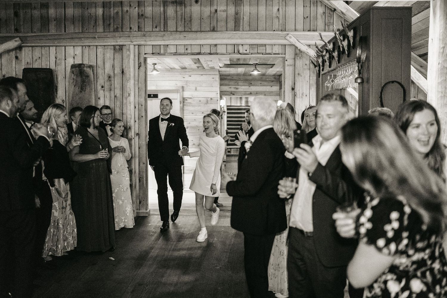 Newlyweds make grand entrance into rustic barn wedding reception as guests applaud in black and white photo.
