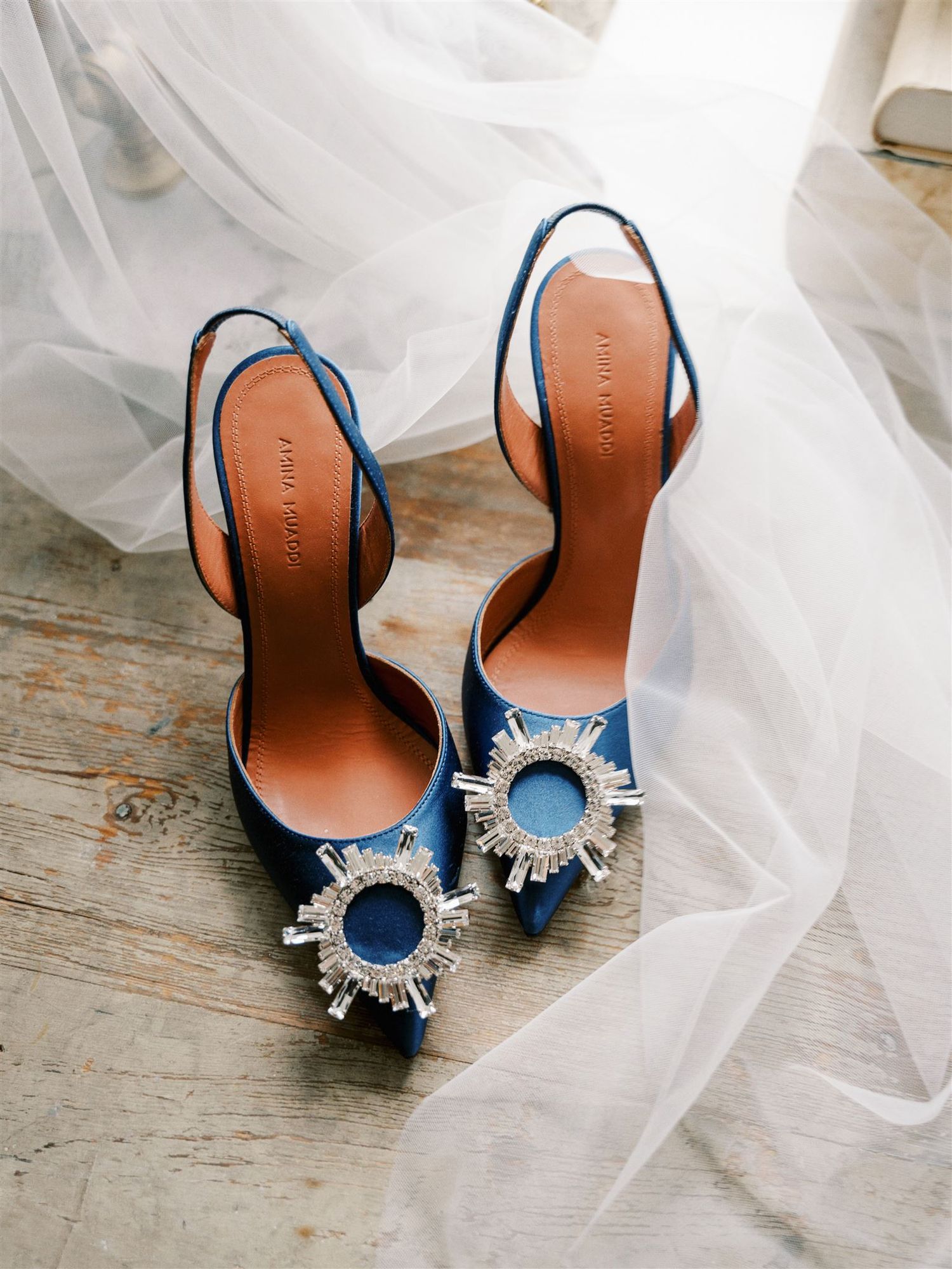 Blue satin bridal heels with crystal sunburst embellishments resting on wood floor with veil.