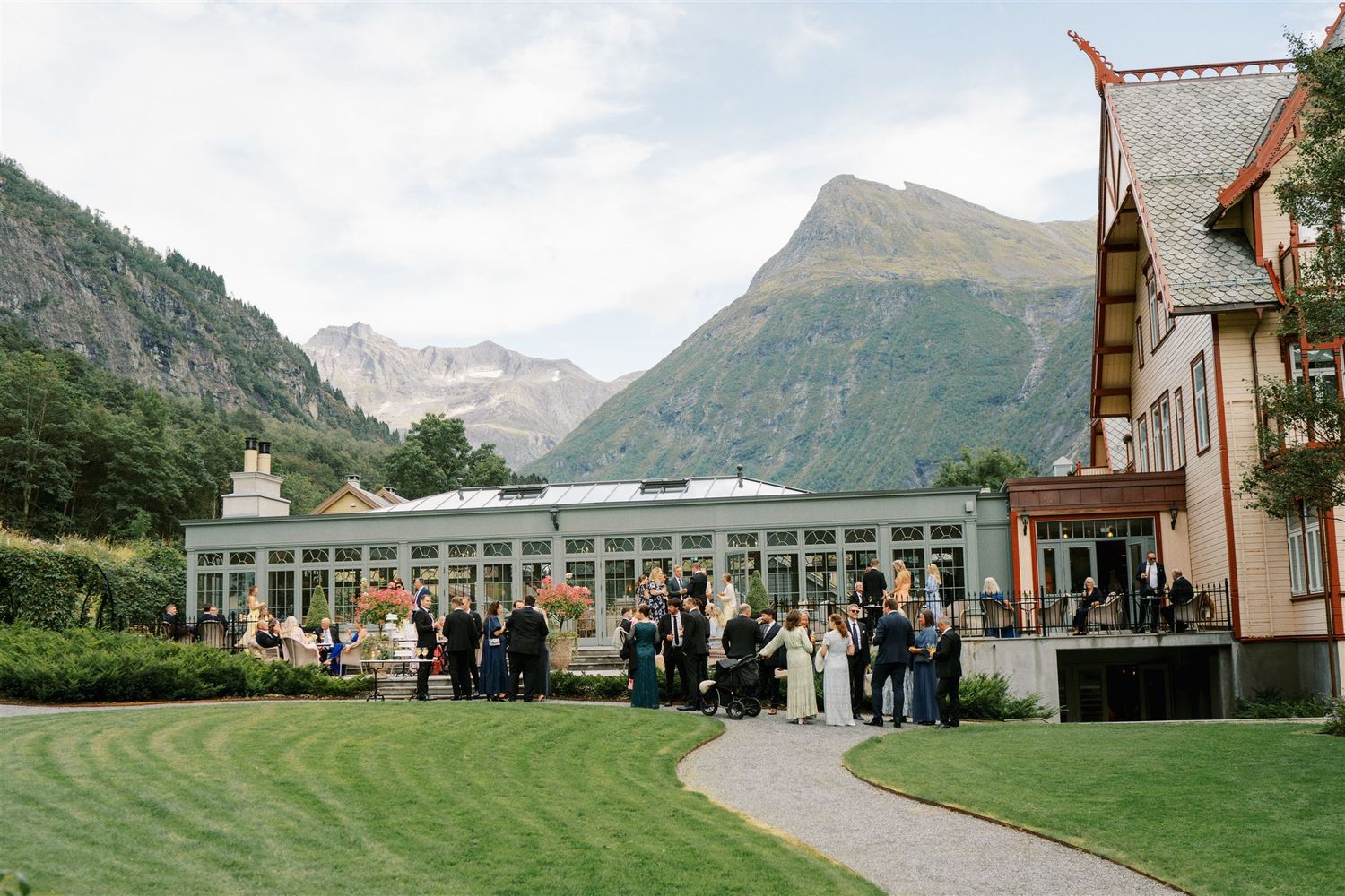 Elegant wedding reception venue with mountain backdrop, guests mingling on green lawn beside glass conservatory.