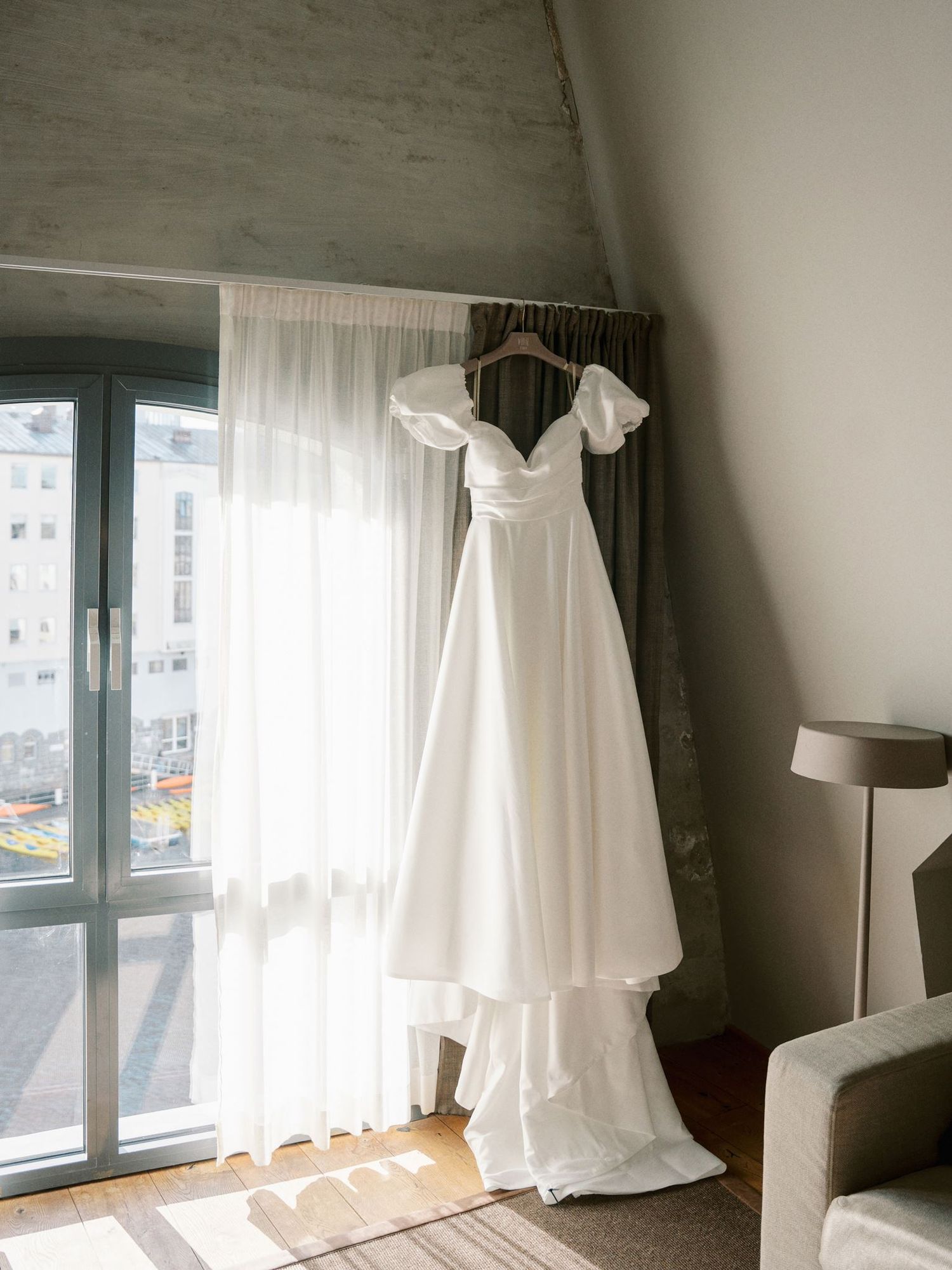 White A-line wedding dress with puff sleeves hanging by a sunlit window in a modern hotel bridal suite.