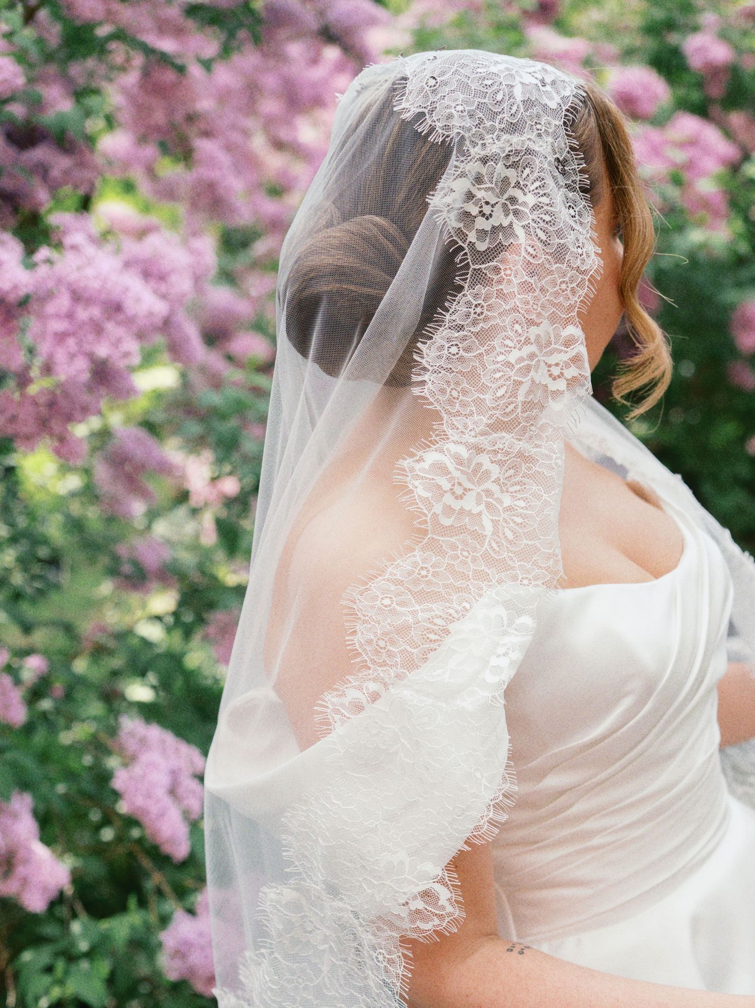 Bride wearing lace-trimmed veil and strapless gown, posed before blooming pink lilac bushes.