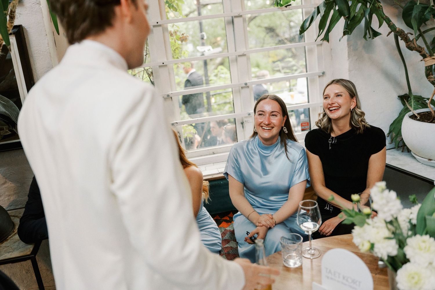 Two women smiling and laughing at a dinner table with wine glasses and white flowers in a bright restaurant.