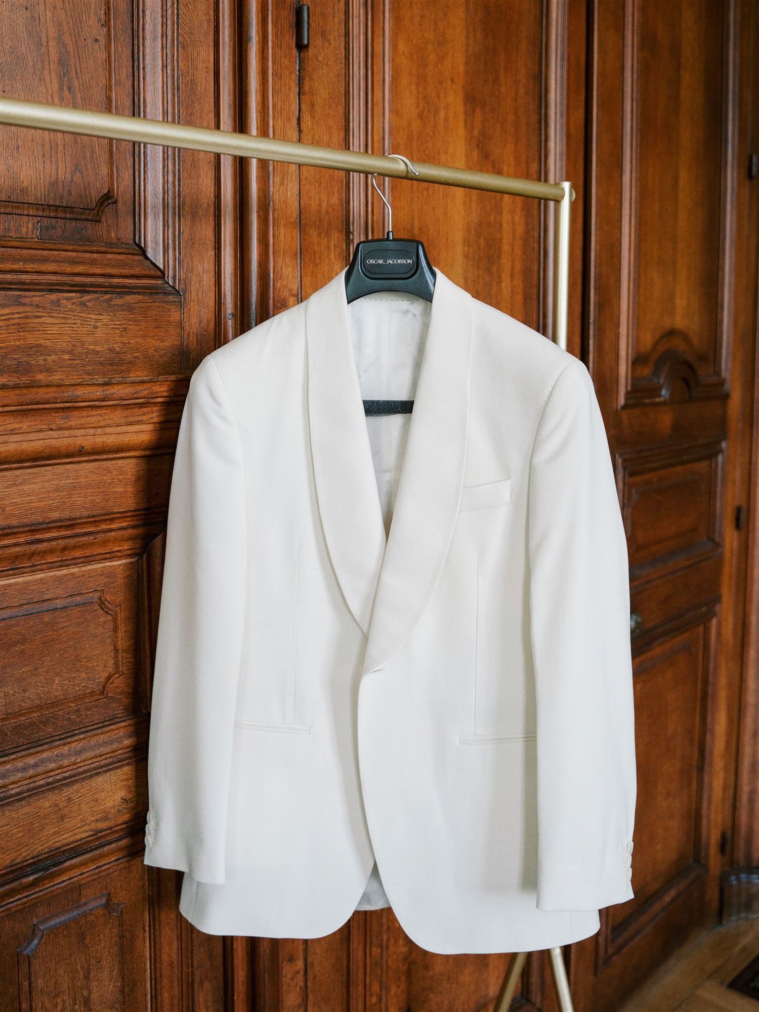 White shawl lapel tuxedo jacket hanging on brass rail against rich dark wood paneled wall.