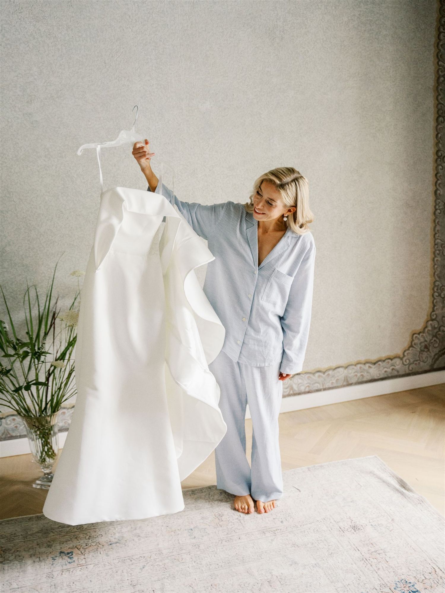 Joyful bride in blue pajamas holding up her elegant ruffled white wedding gown on a hanger.