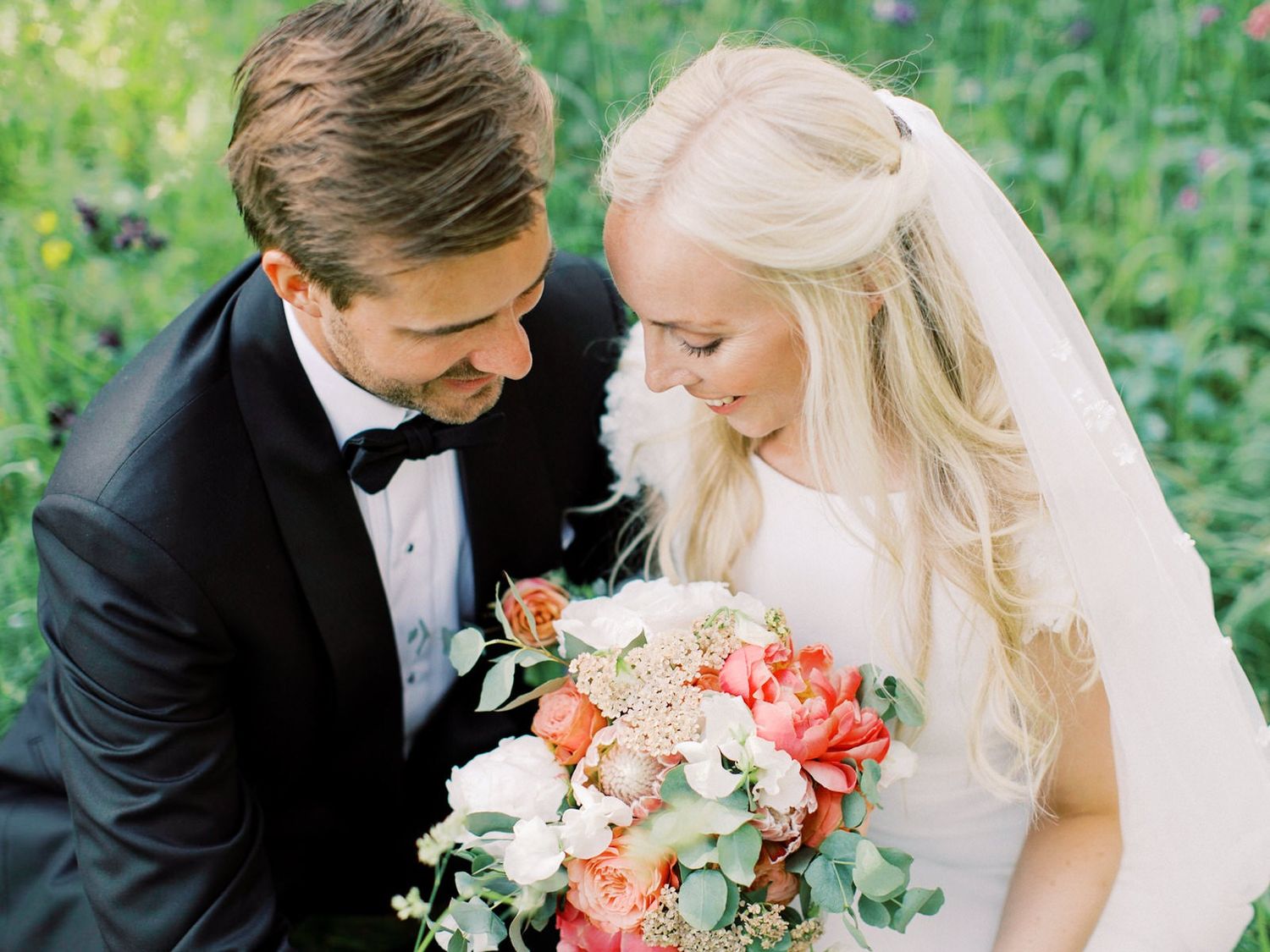 Smiling bride and groom in tuxedo sharing romantic moment over colorful coral bridal bouquet.