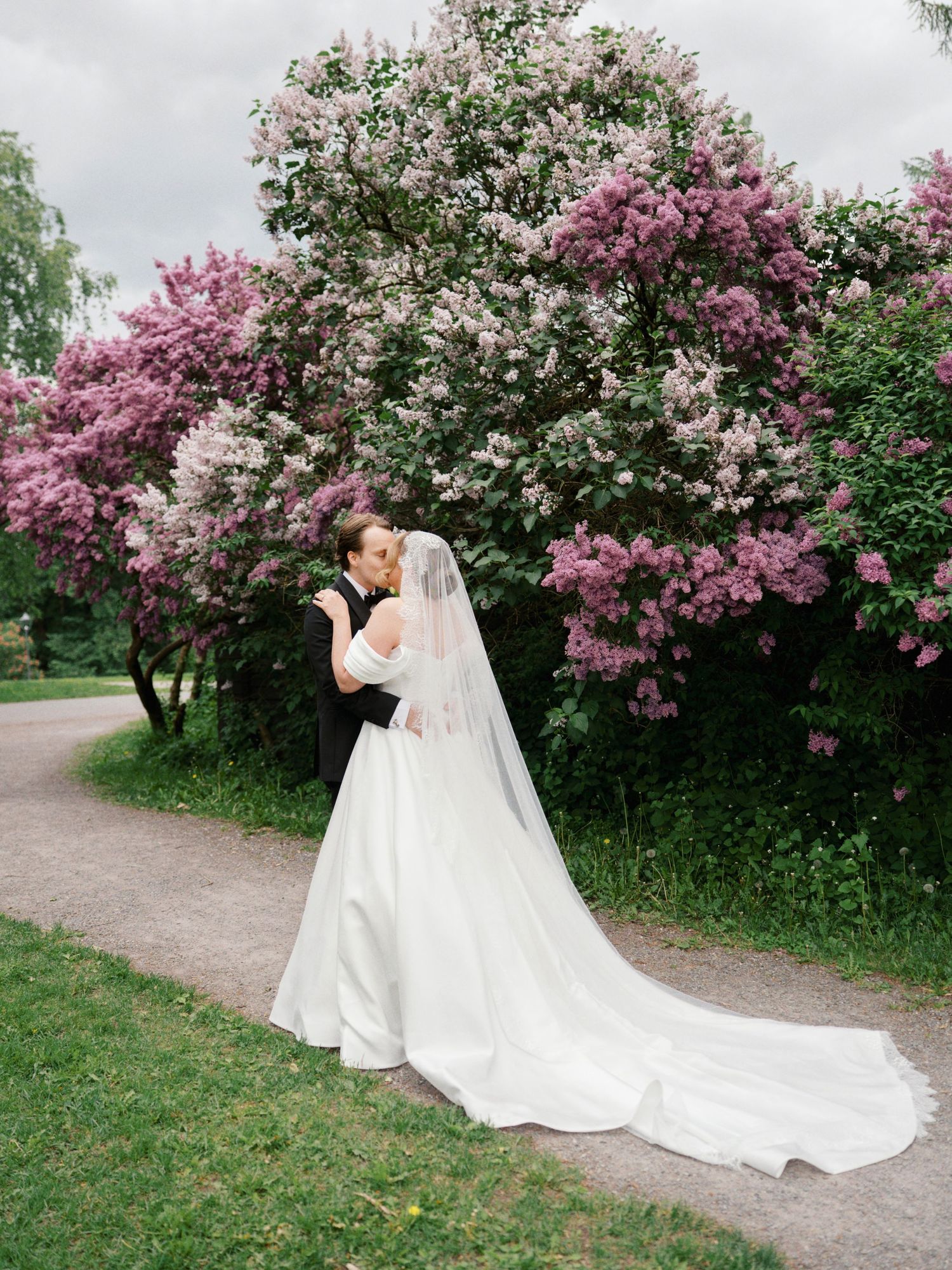 A bride and groom share a romantic kiss in front of blooming pink and white lilac trees on their wedding day.