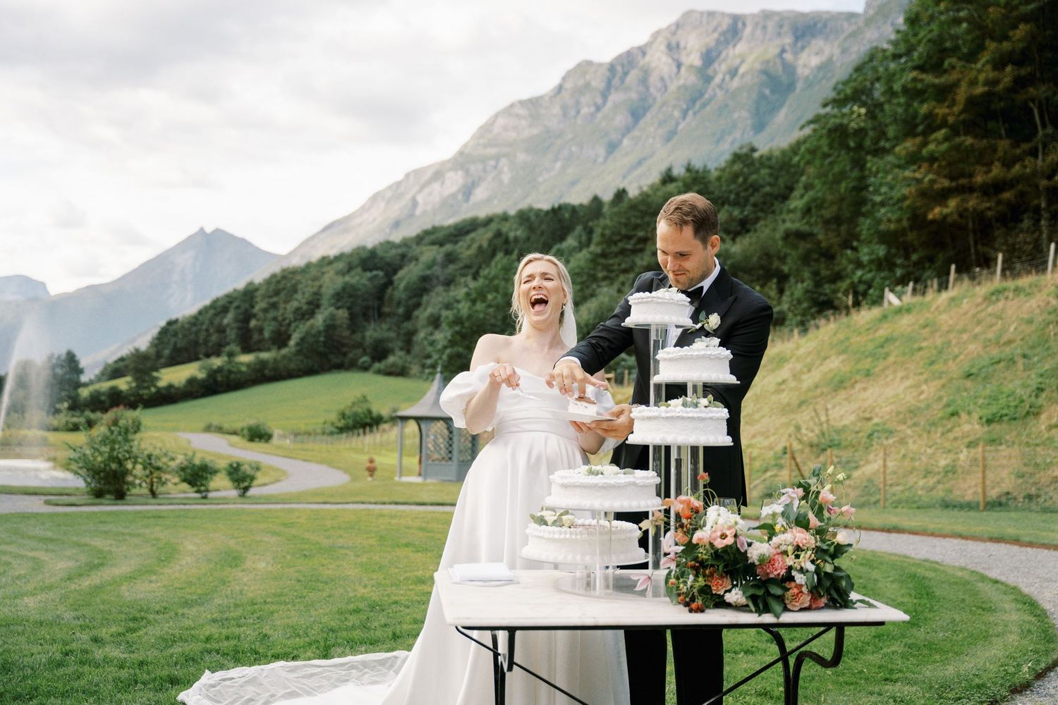 Bride and groom cutting a tiered wedding cake outdoors with stunning mountain and green valley backdrop.