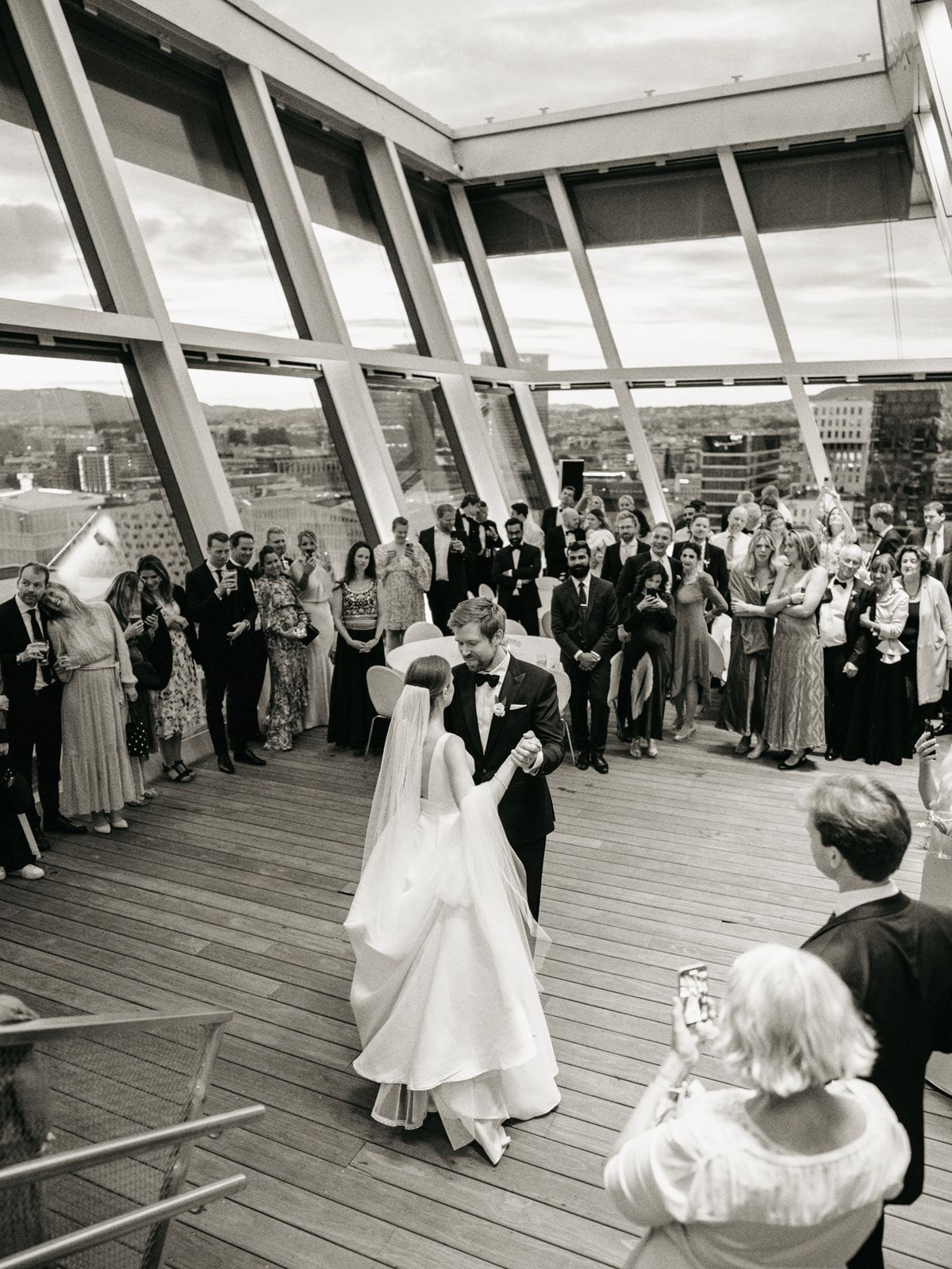 Bride and groom sharing their first dance on a rooftop surrounded by wedding guests in black and white.