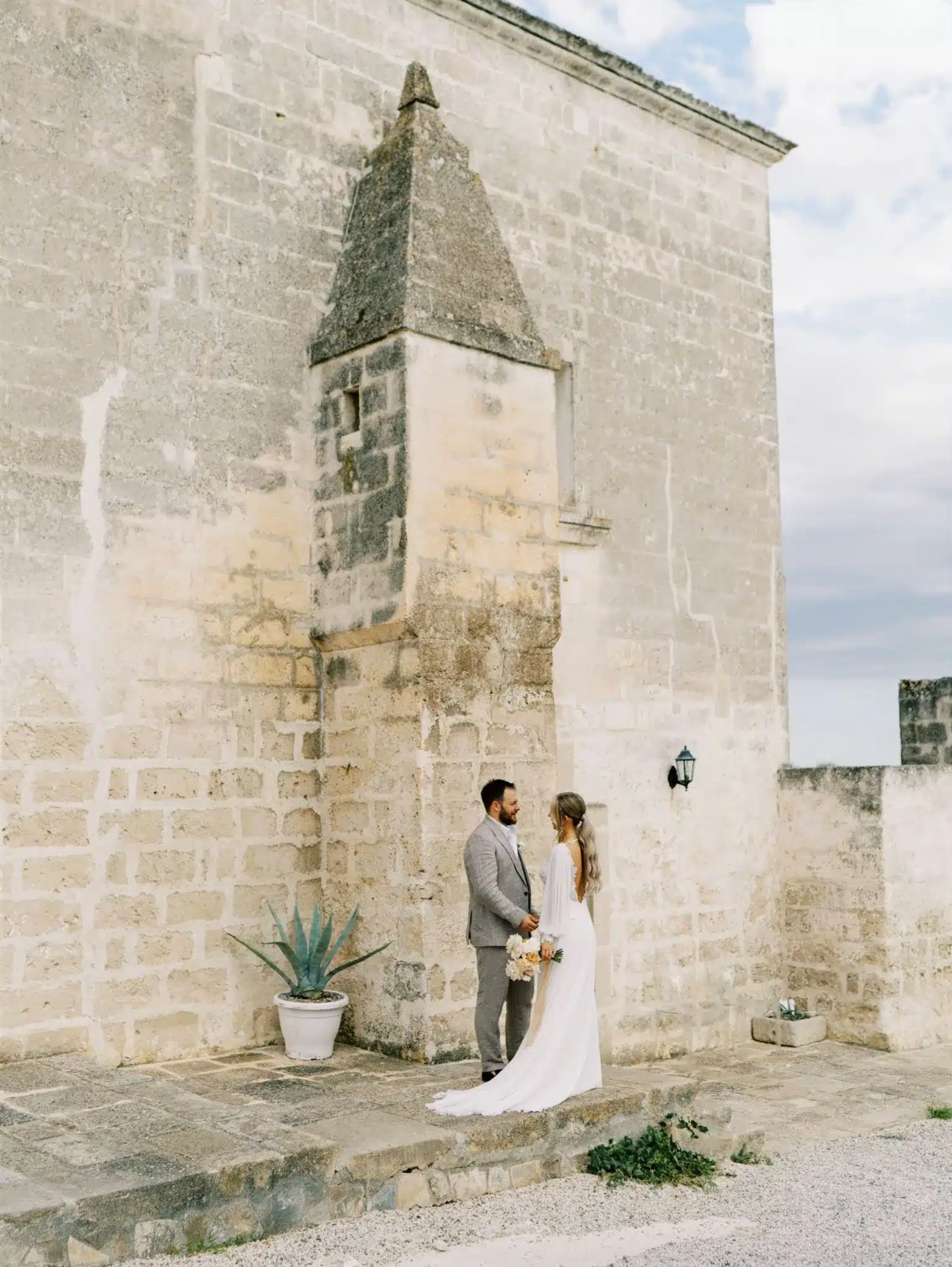 Bride and groom in elegant attire stand together beside ancient stone building in Italy.