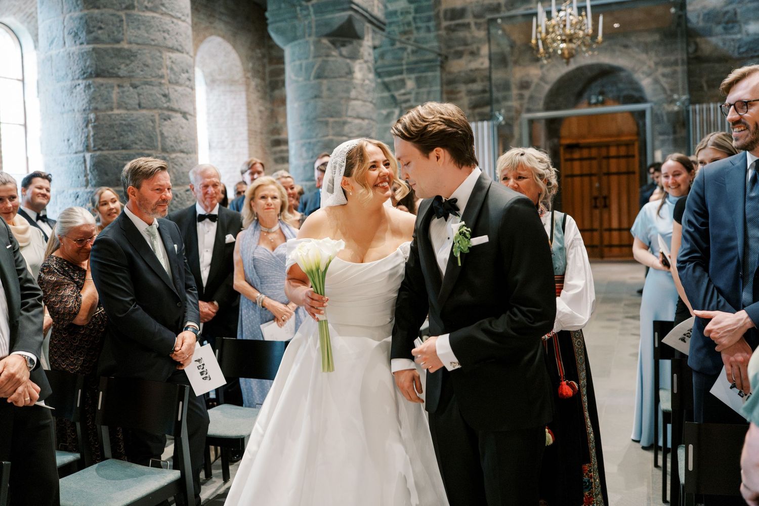 Bride and groom share a joyful moment walking down the aisle in a historic stone church surrounded by wedding guests.