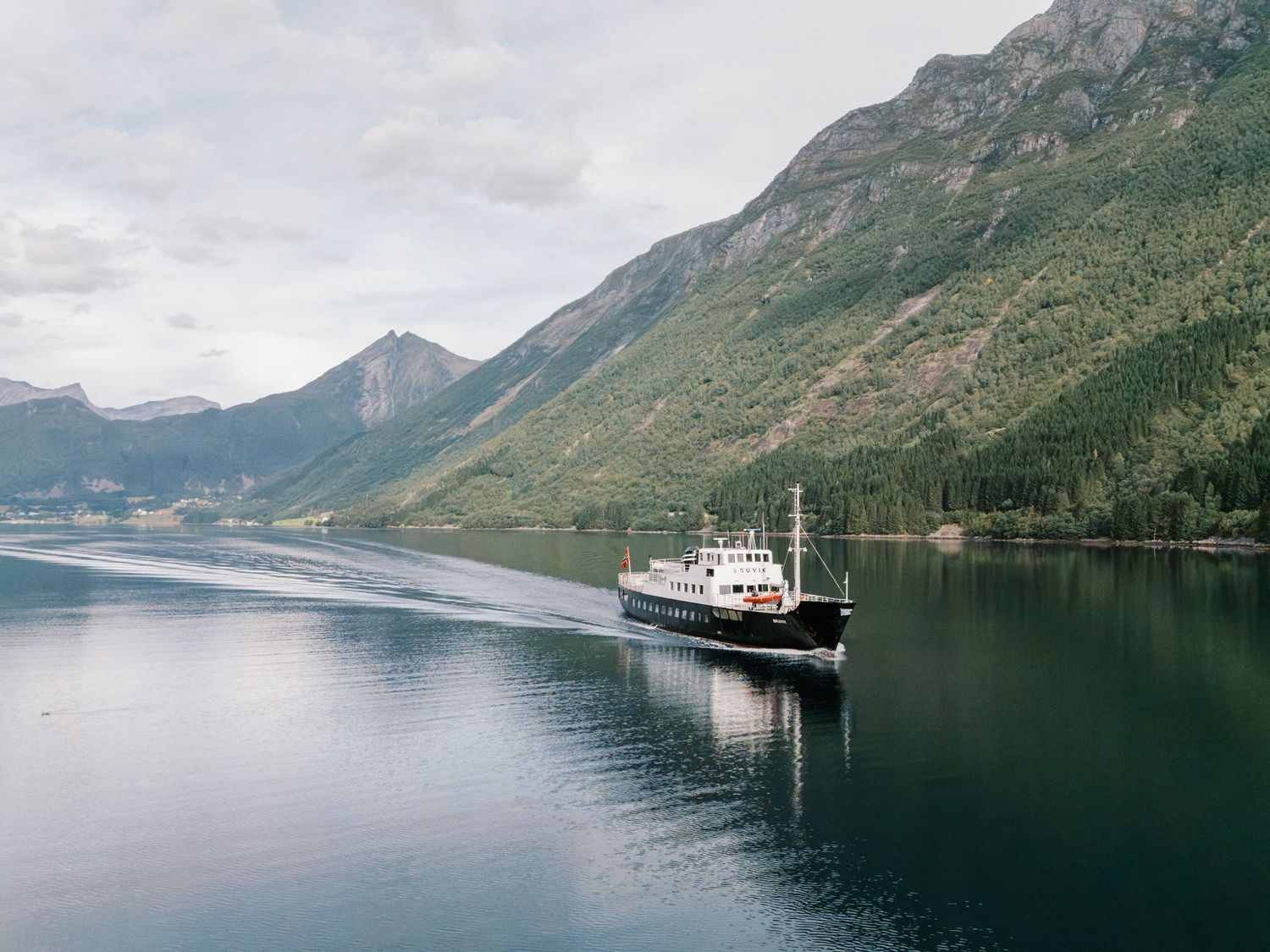 A ferry boat cruises through a calm Norwegian fjord surrounded by lush green mountains under a cloudy sky.